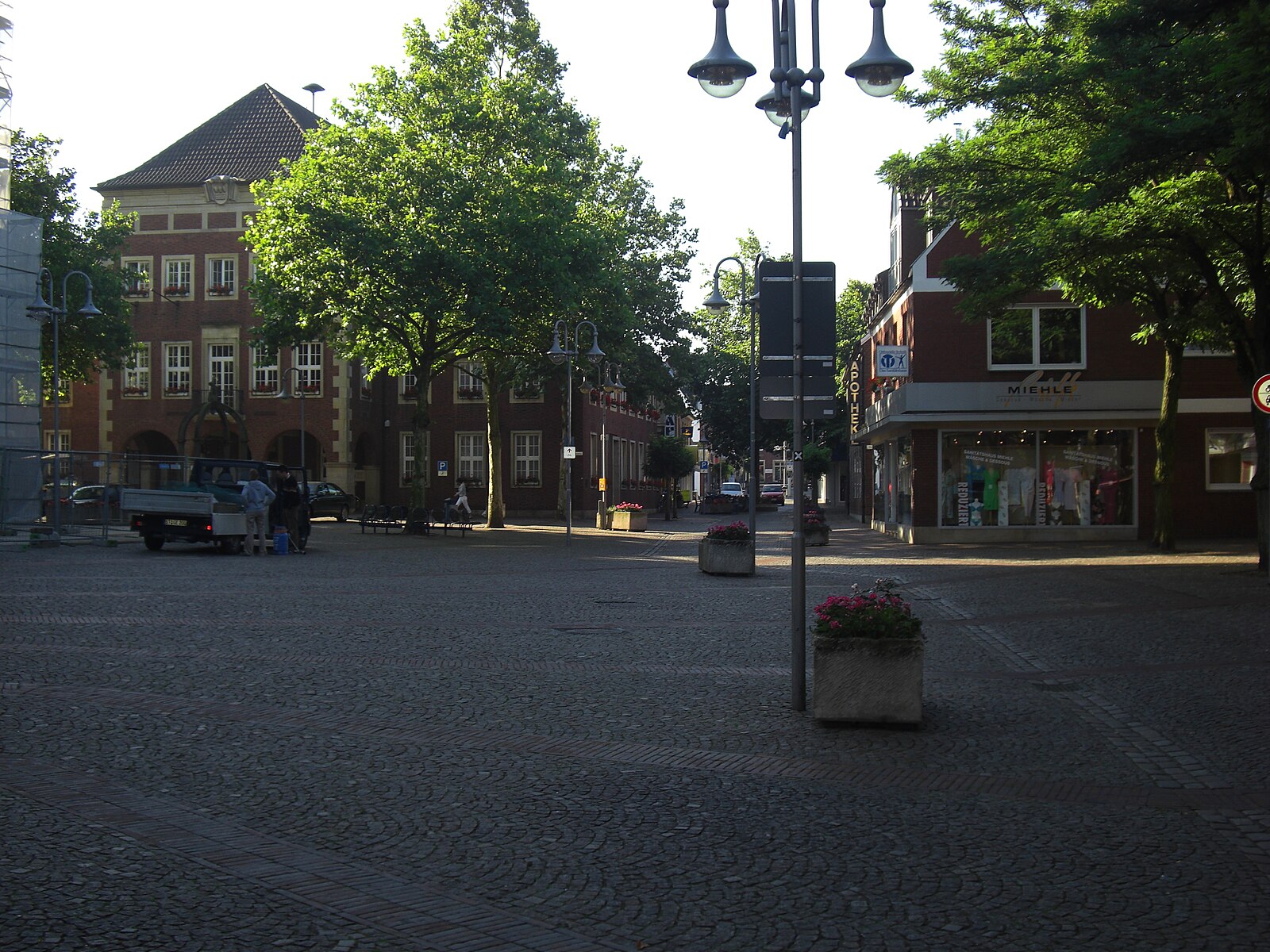 Blick auf den Marktplatz in Stadtlohn mit Kopfsteinpflaster, Blumenbeeten, Geschäften und historischen Backsteingebäuden im Abendlicht.
