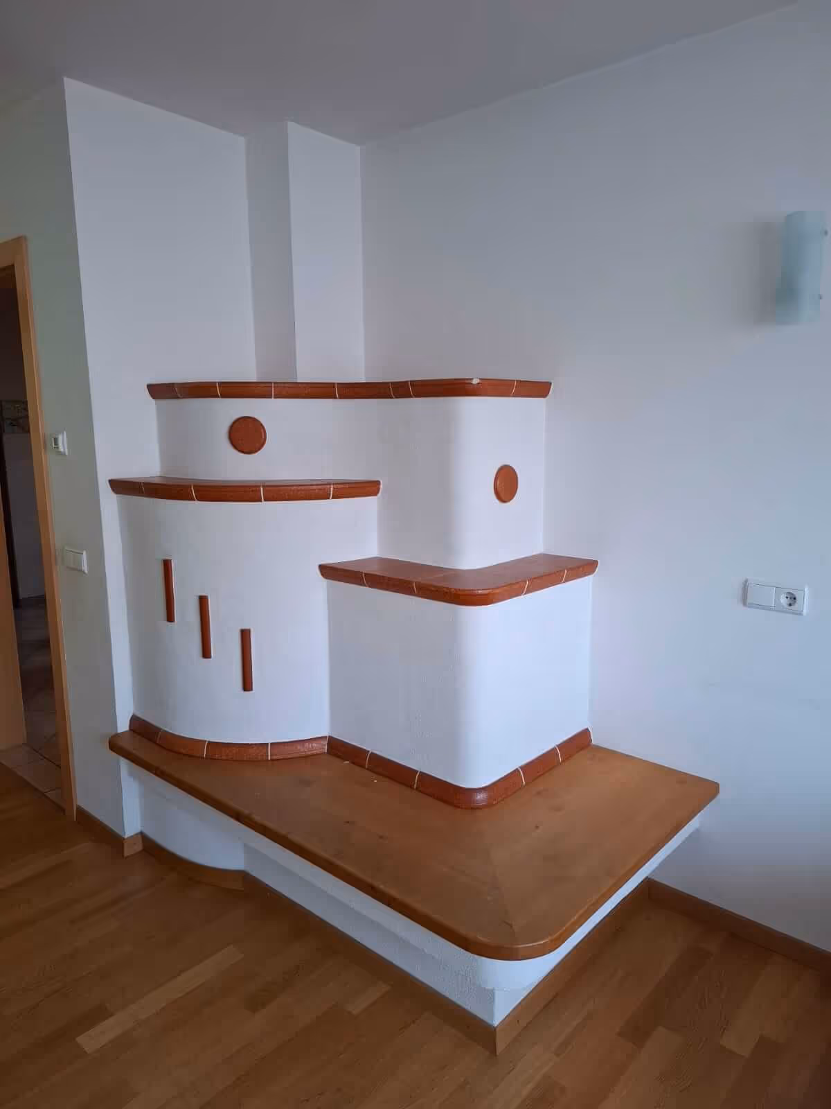 Stufig aufgebauter Speicherofen mit verputzter Oberfläche, roten Keramikelementen und seitlicher Holzbank.