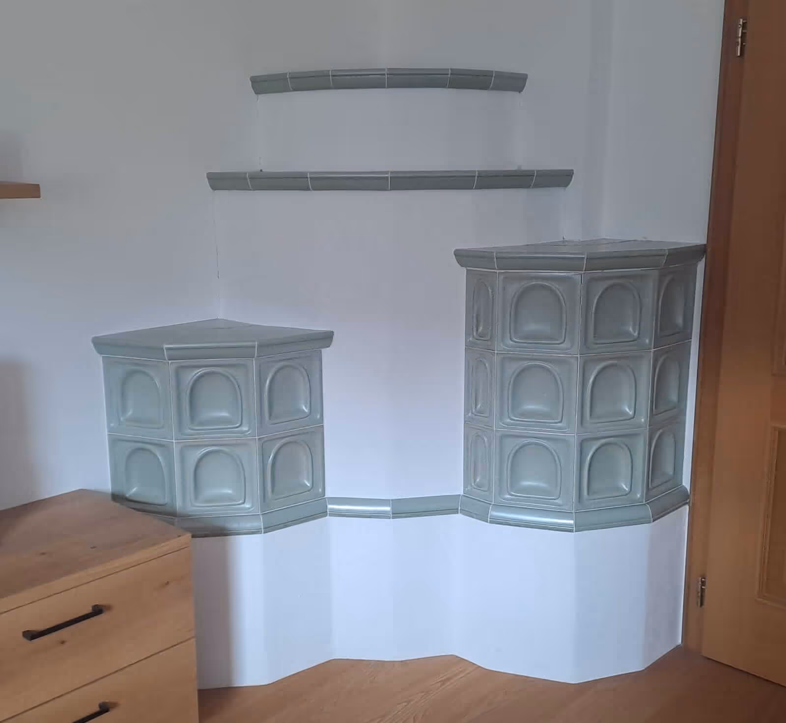 Traditioneller grün glasierter Kachelofen in einer Zimmerecke mit zwei unterschiedlich hohen Ofenteilen und halbrunden Ablagen.
