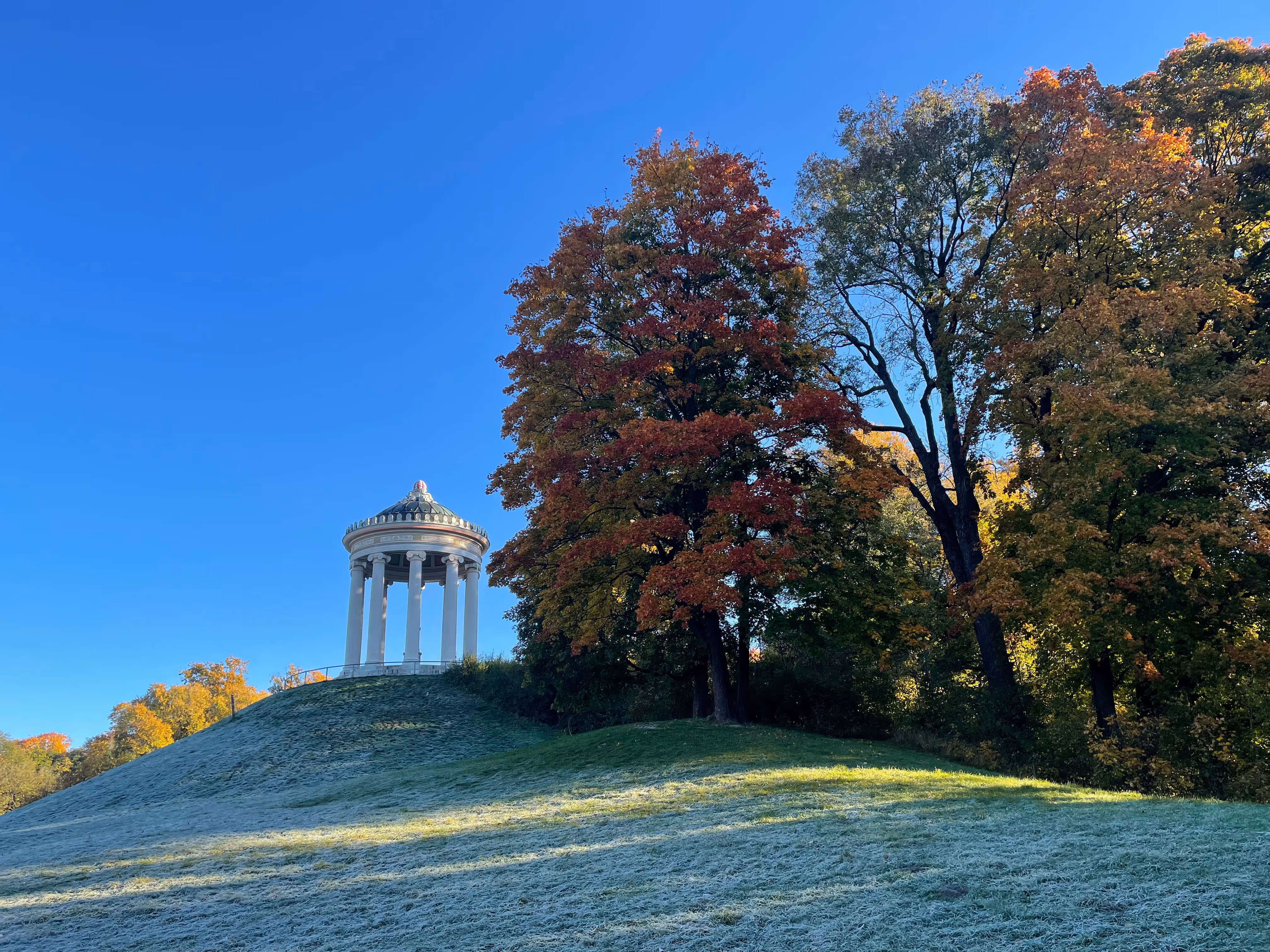 Weißer Pavillon auf einer Anhöhe, von Herbstbäumen umgeben, blauer Himmel.