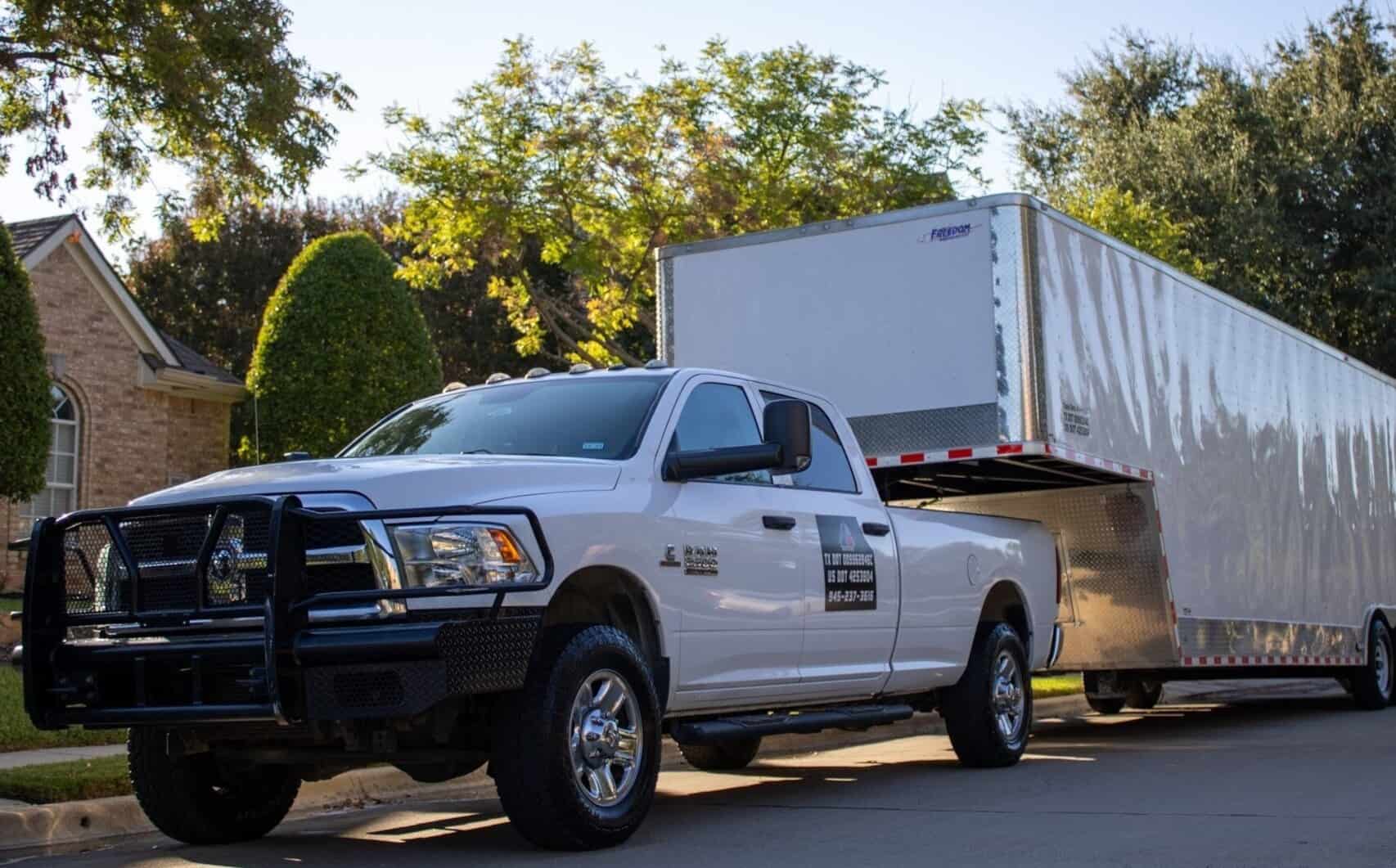 Texas Delta Moving's white truck hitched to a large enclosed trailer parked in front of a suburban home. The truck is branded with the company logo and contact information, emphasizing professionalism and reliable moving services. The well-maintained landscaping and bright sunlight highlight the residential setting, making it clear that Texas Delta Moving specializes in residential moves. The powerful truck and spacious trailer convey the company's ability to handle large-scale moves efficiently.