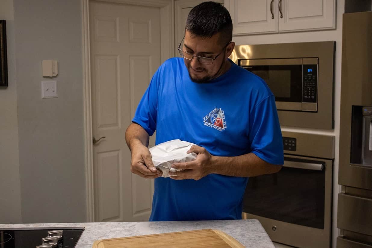 Texas Delta Moving team member carefully packing a kitchen item in a customer's home. The mover, wearing a branded blue company shirt, wraps a fragile object with protective paper, ensuring it is securely prepared for transport. The modern kitchen setting, complete with stainless steel appliances, emphasizes the professional and attentive service Texas Delta Moving provides when handling delicate household items.