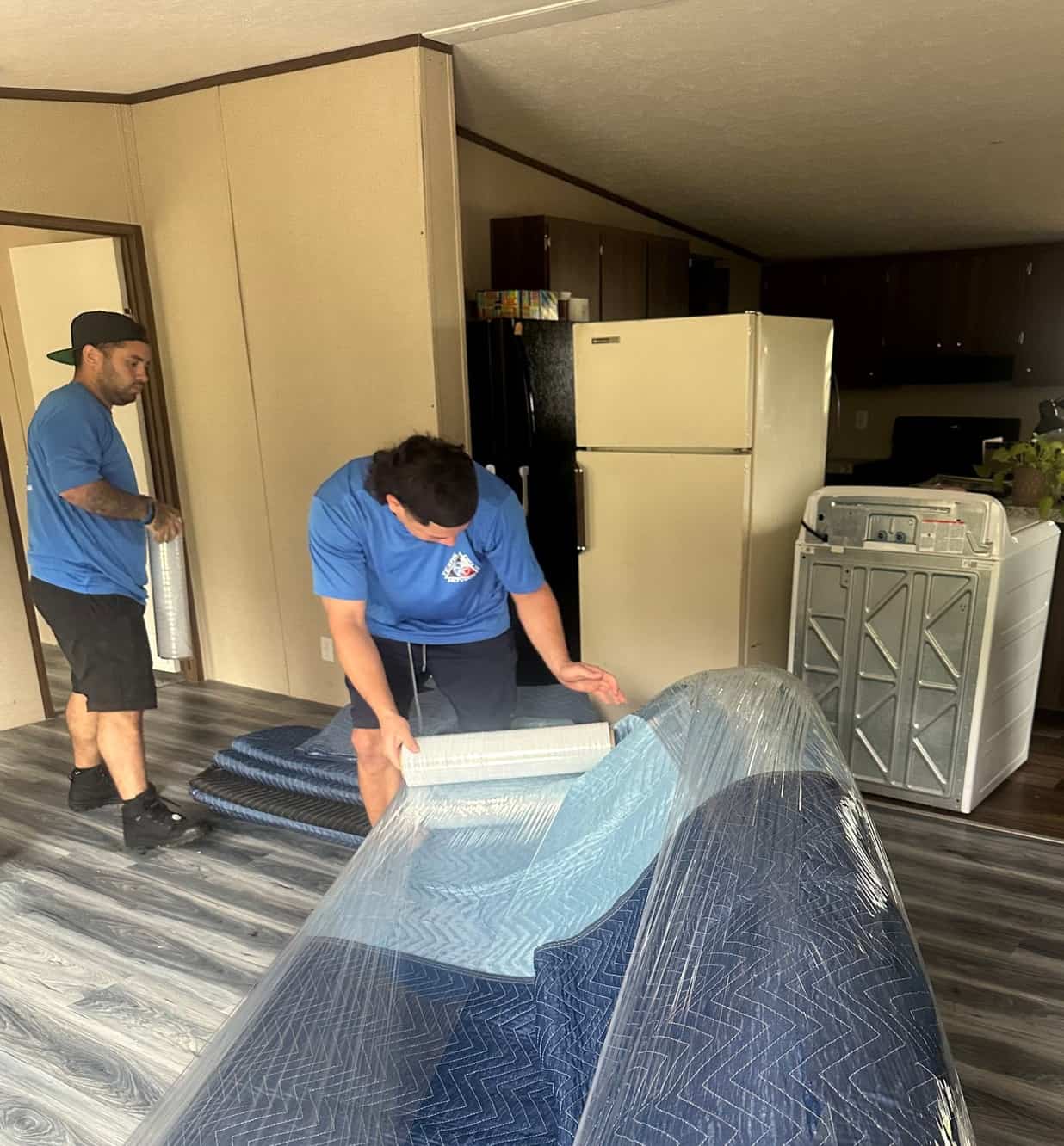 Texas Delta Moving team wrapping a large piece of furniture in protective blankets and plastic wrap during a residential move. The movers, dressed in blue company-branded shirts, are ensuring the item is well-protected for transport. The room is part of a lived-in home, with a refrigerator and other appliances visible in the background. This image highlights the company's focus on careful packing and attention to detail when moving household goods.