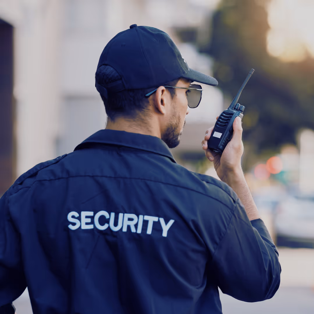 A uniformed security guard using a walkie-talkie while monitoring an outdoor area, wearing a black cap and sunglasses.