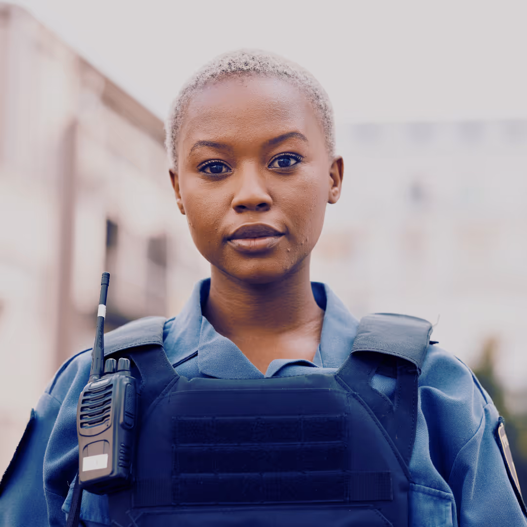 A female security officer in uniform and tactical vest, facing forward with a serious expression, wearing a shoulder radio and standing outdoors.