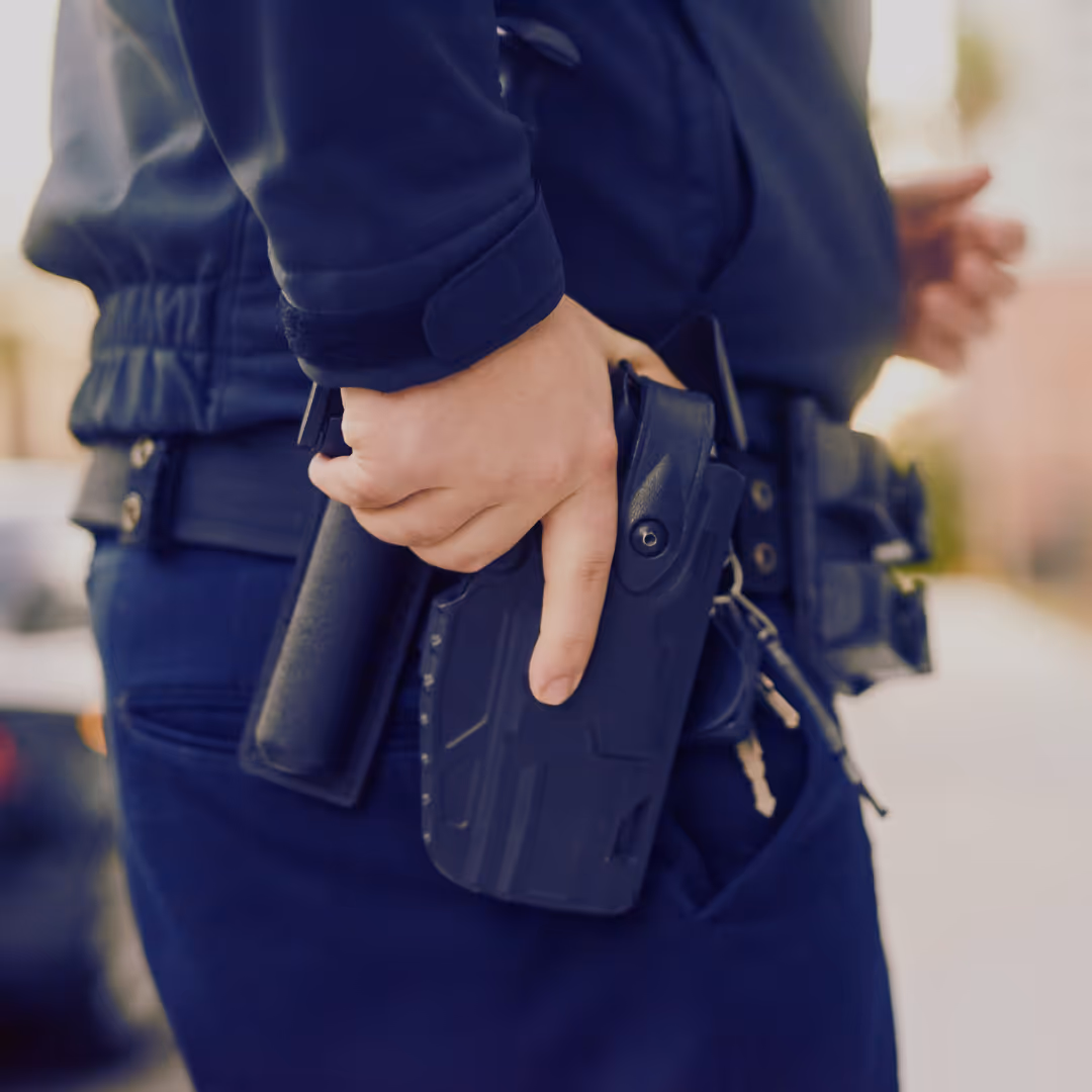A close-up of a security officer’s duty belt, showing a holstered firearm and tactical gear, with the officer’s hand resting on the weapon.