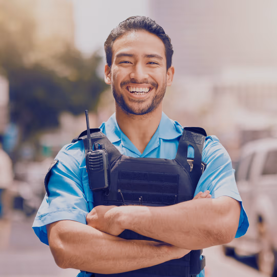 A smiling male security officer in uniform and tactical vest, standing confidently with arms crossed on a city street, wearing a shoulder radio.
