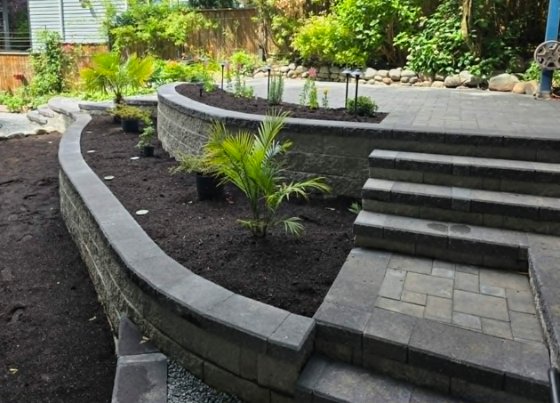 Curved stone retaining wall with planted soil areas and stone steps leading to a paved patio.