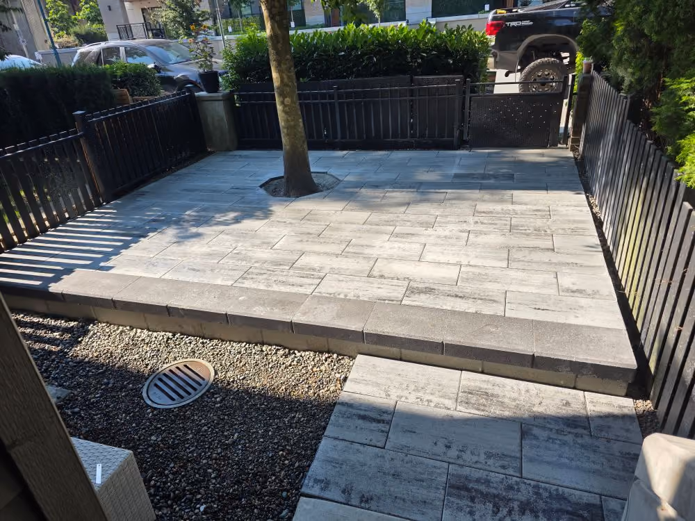 Backyard patio with gray stone tiles, a tree planted in the center, and black metal fencing around.