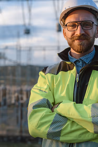 Man in safety gear and hard hat standing outdoors, smiling confidently.