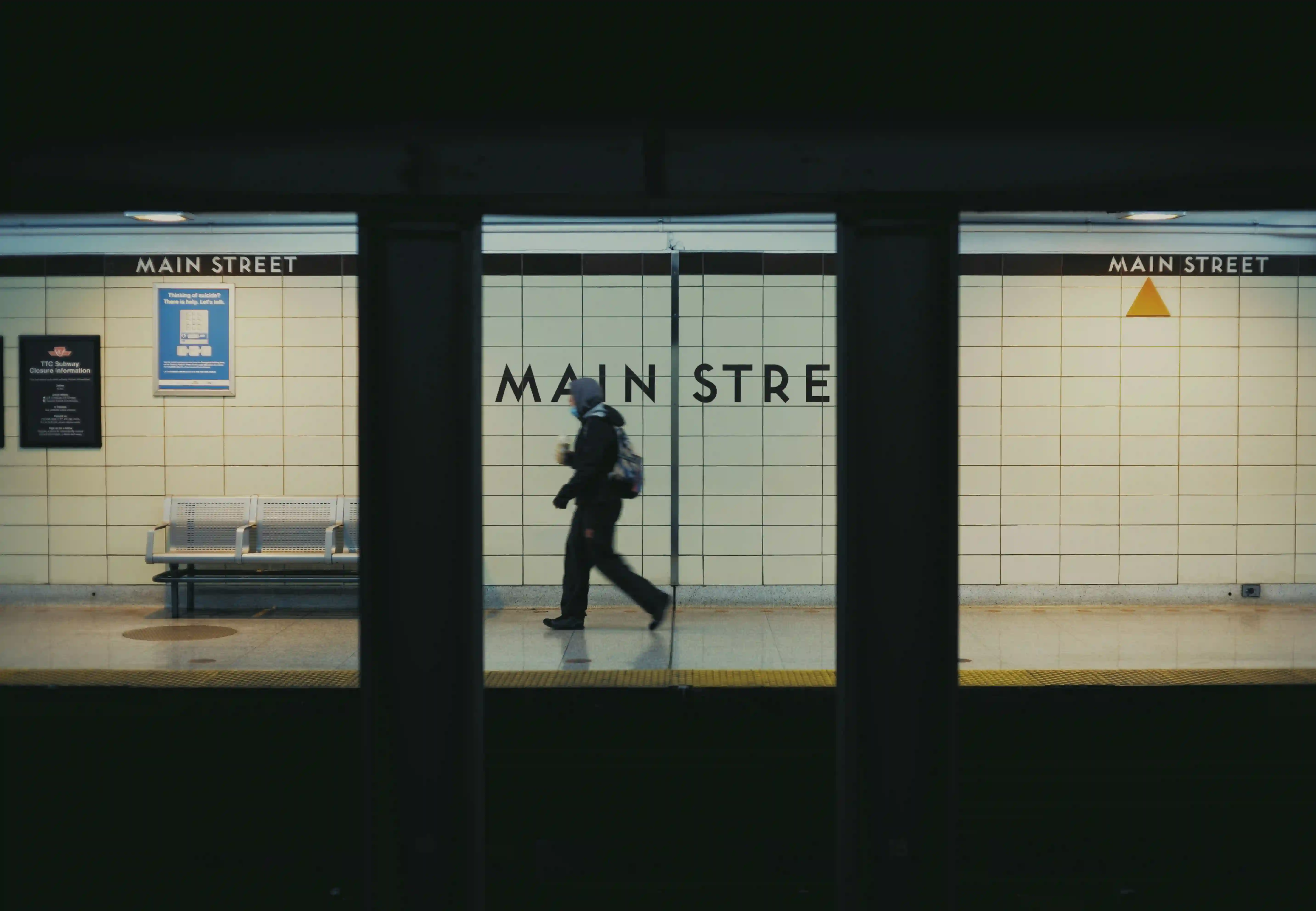 Person wearing a hood and mask walking on the Main Street subway platform with tiled walls and benches.