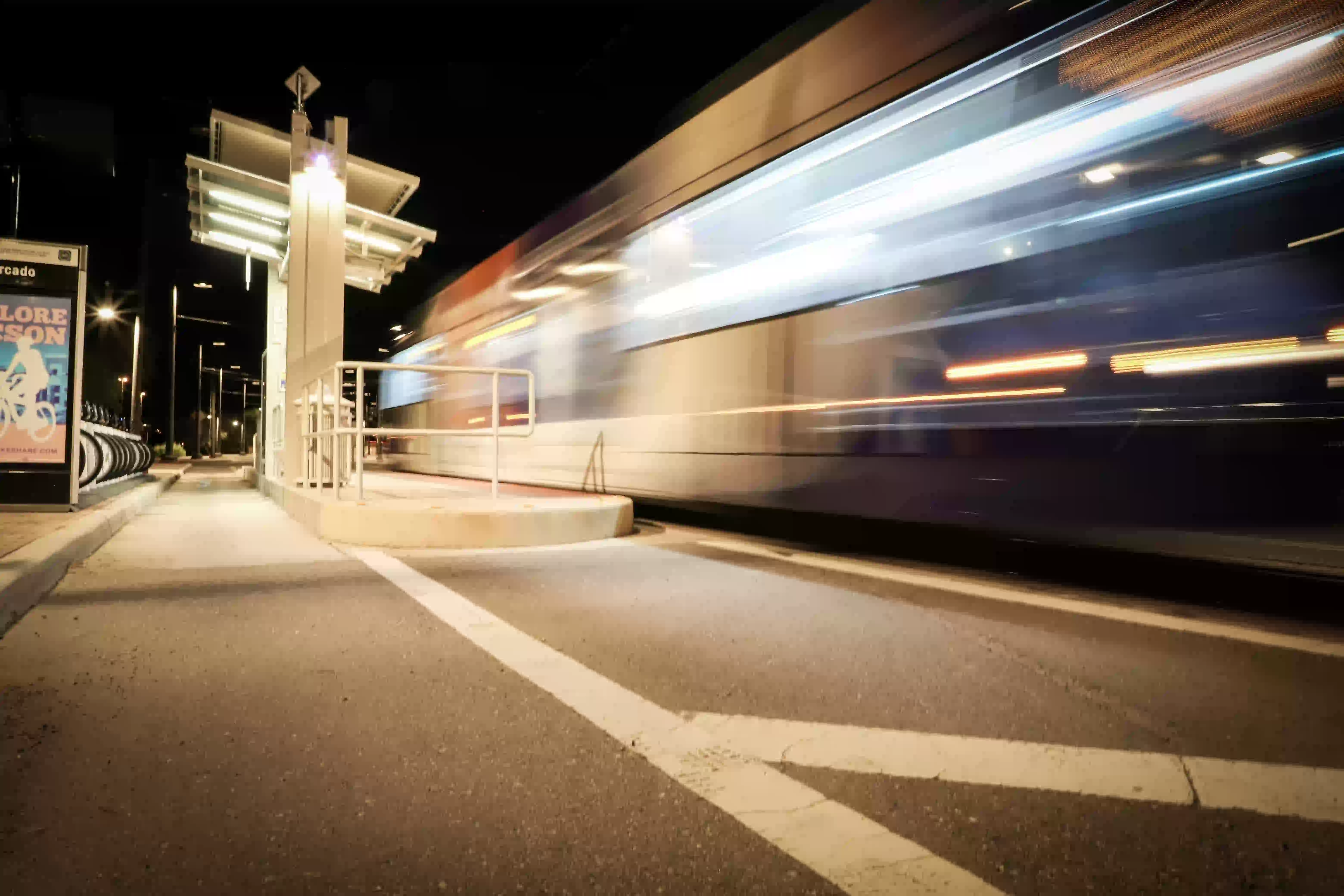 Blurred light streaks of a train passing by a well-lit train station platform at night.