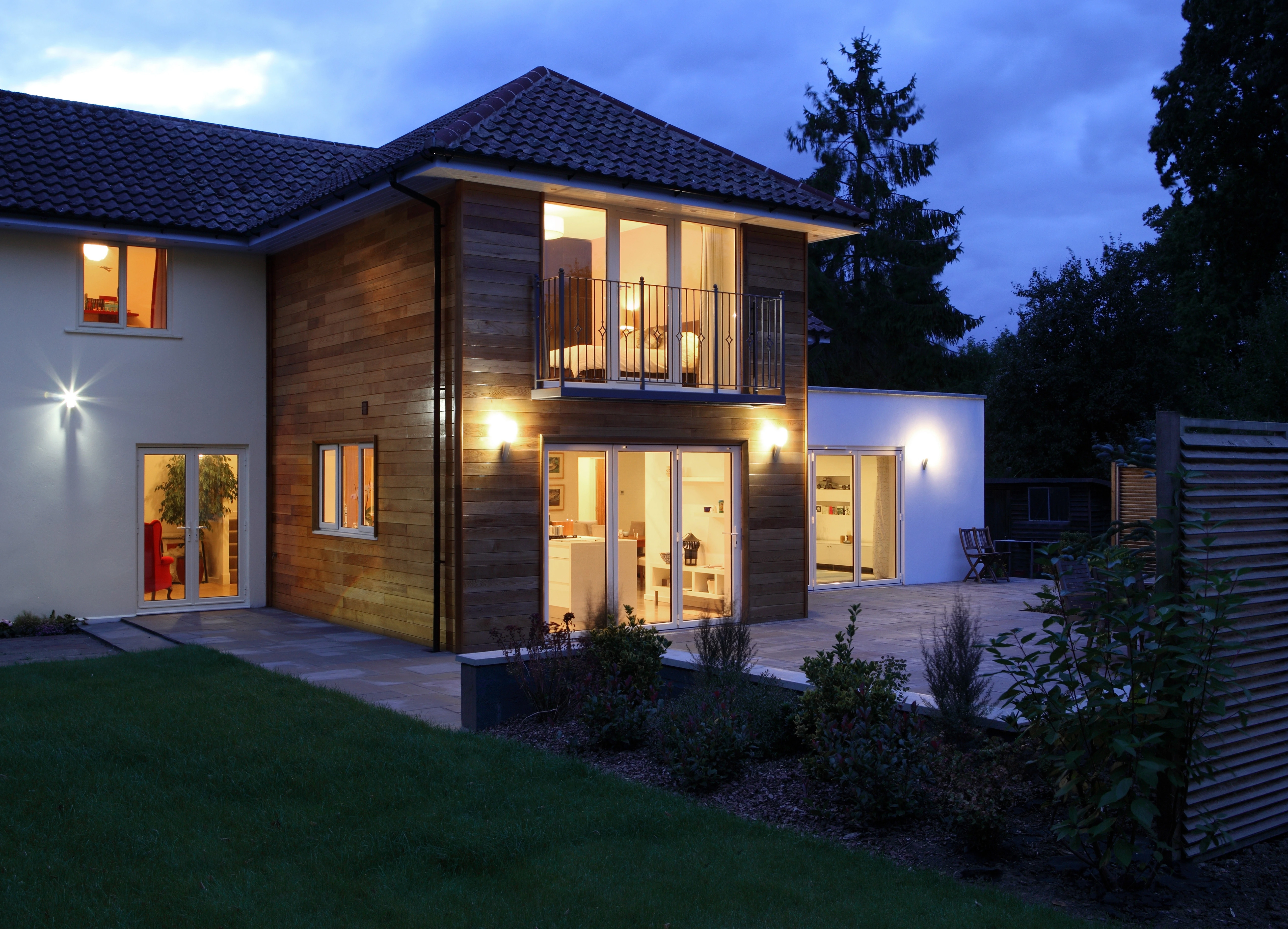 Modern two-story house at dusk with warm interior lights, a wooden facade section with balcony, large glass doors opening to a tiled patio, green lawn, and garden with shrubs.