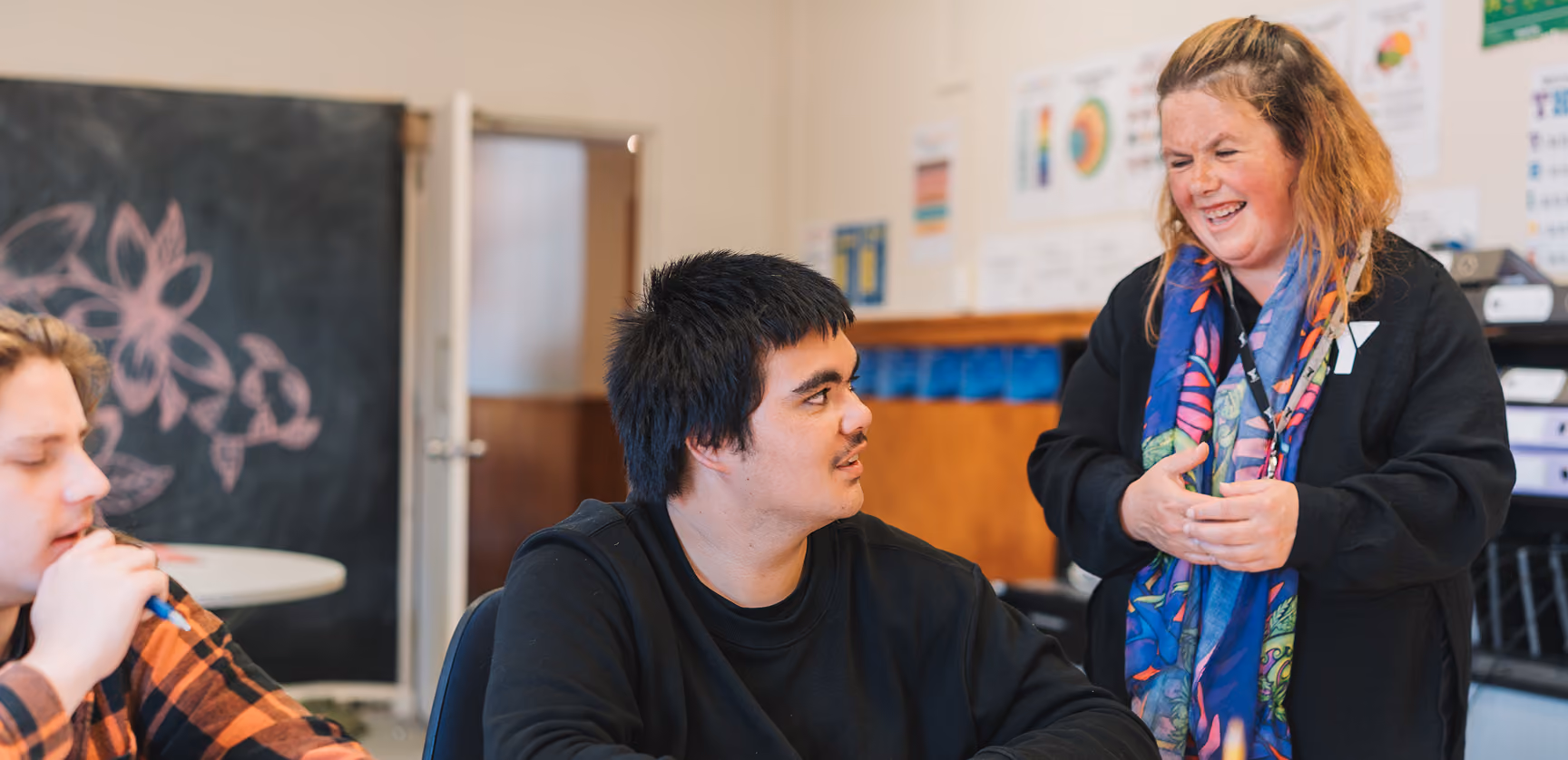 A tutor laughing with a learner in a classroom, with a chalkboard behind them