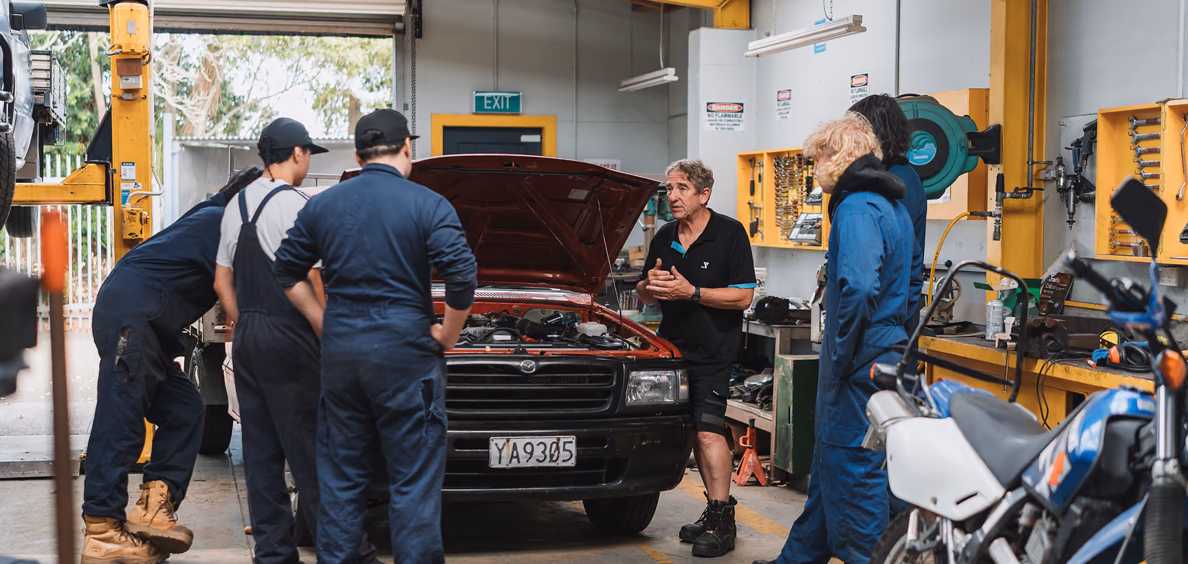 Learners in overalls and a tutor gathered around a car with its bonnet up in an automotive workshop