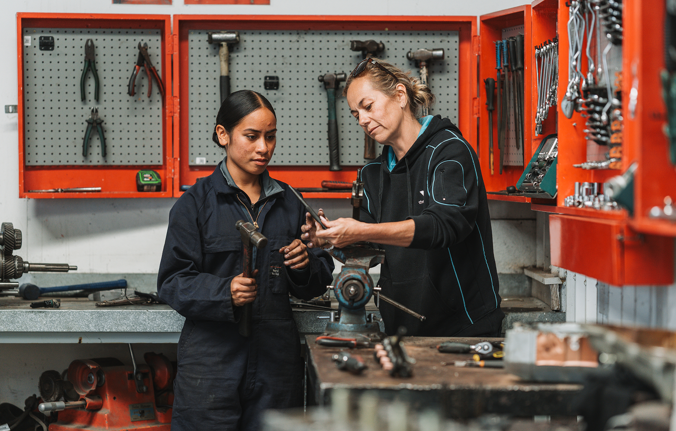 A tutor and a learner working together at a workshop bench, with hand tools hanging on the wall behind them