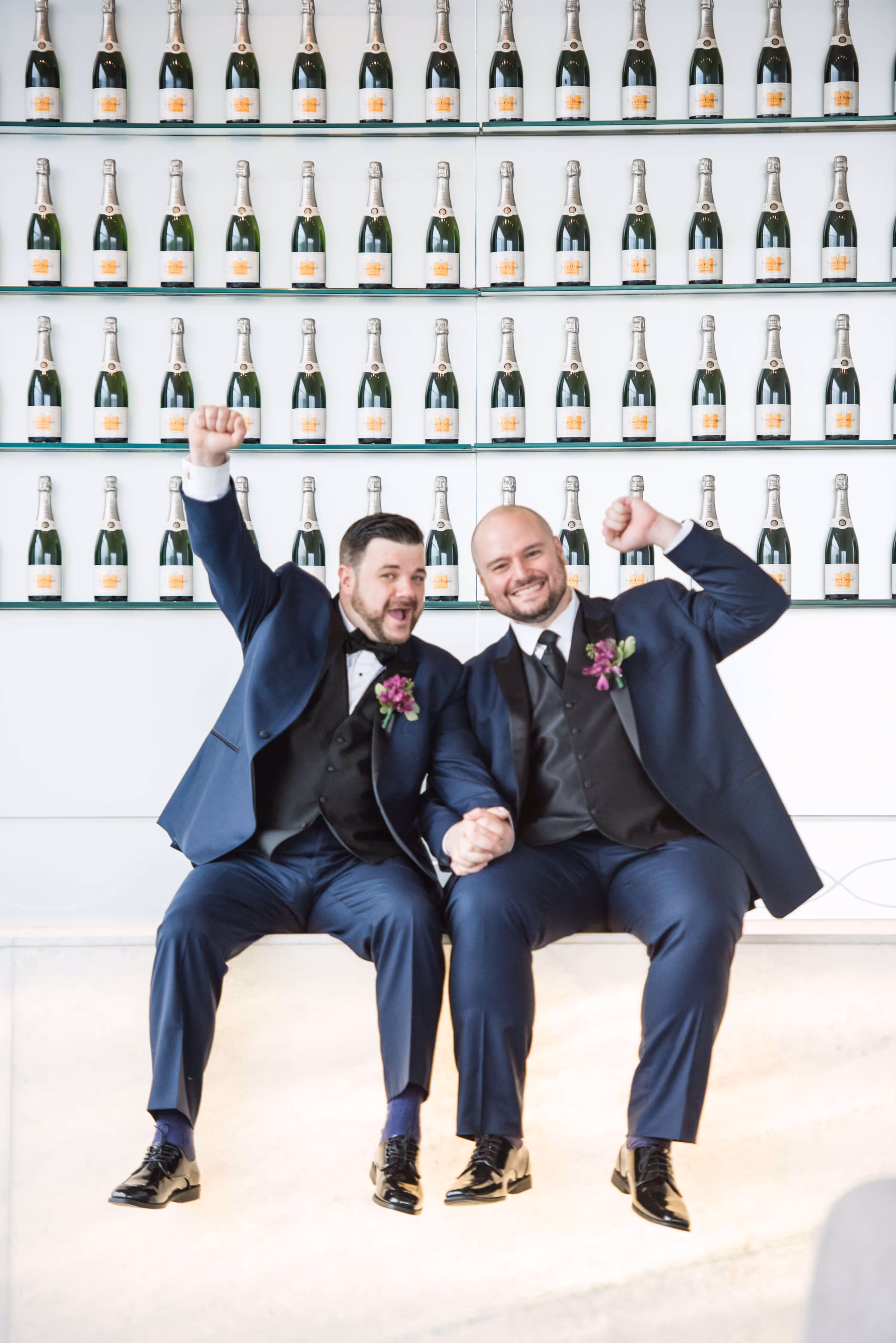Two grooms in navy tuxedos sitting and holding hands with raised fists, smiling in front of shelves filled with champagne bottles.