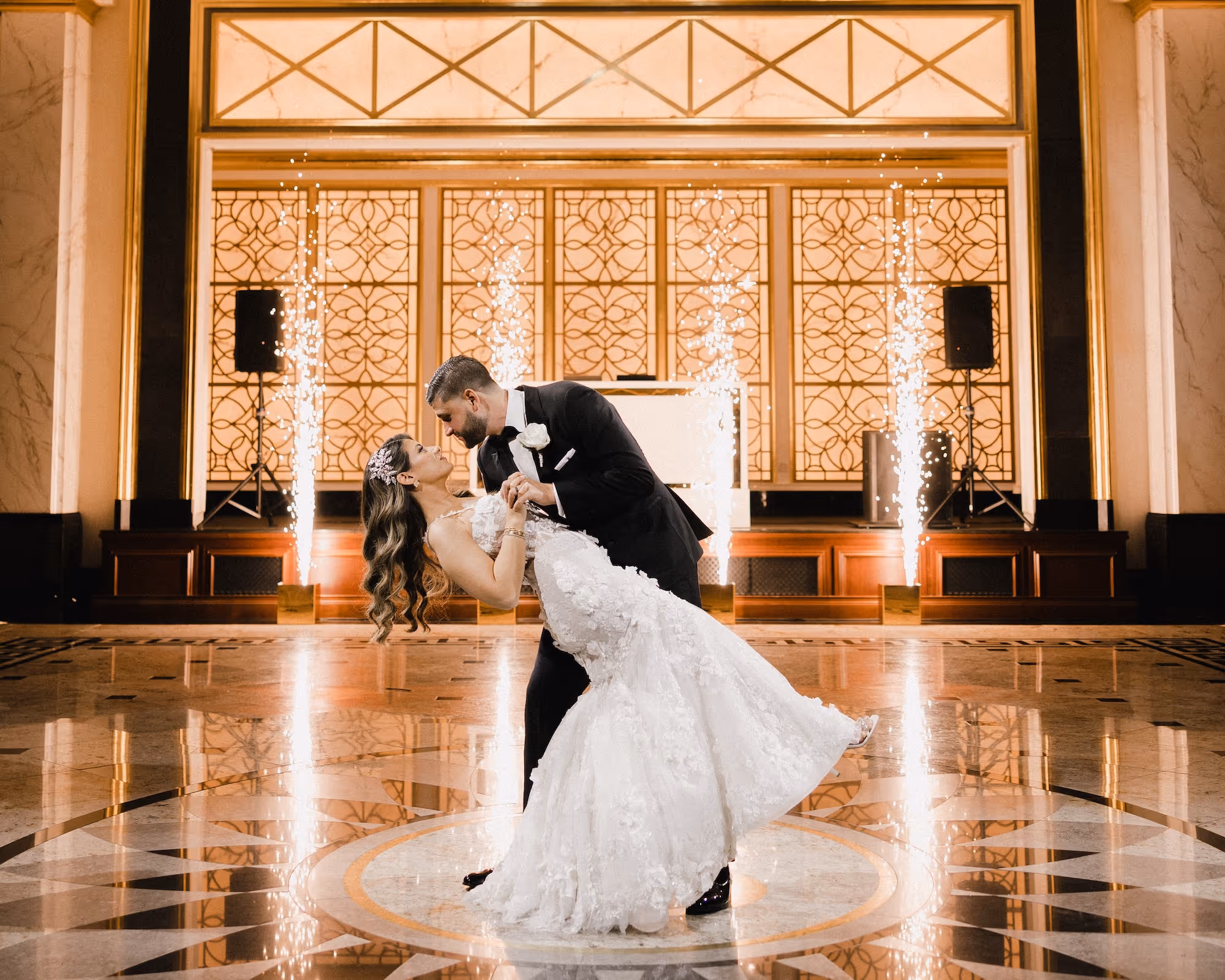 Bride and groom dancing in an elegant hall with fireworks in the background.