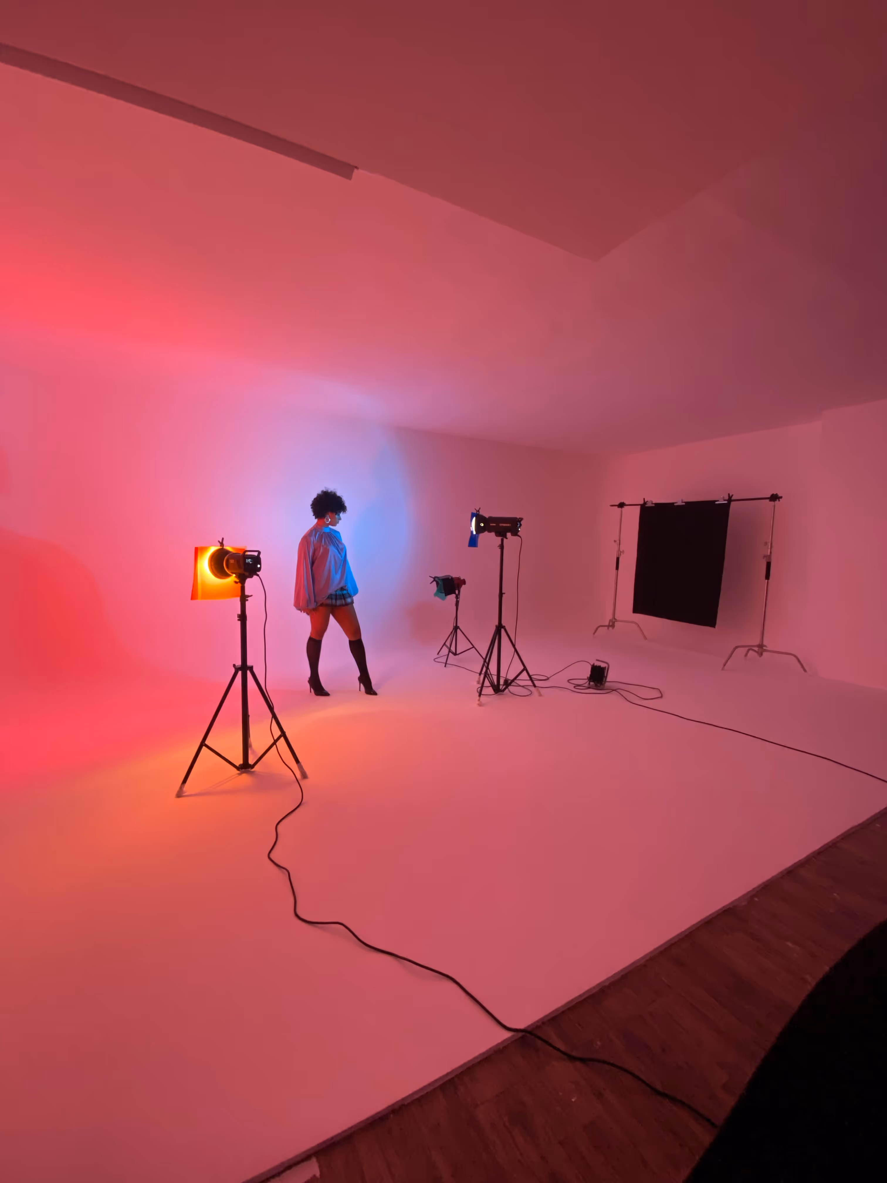 A person posing in a photo studio lit with red and blue lights, surrounded by professional lighting equipment and a black backdrop.