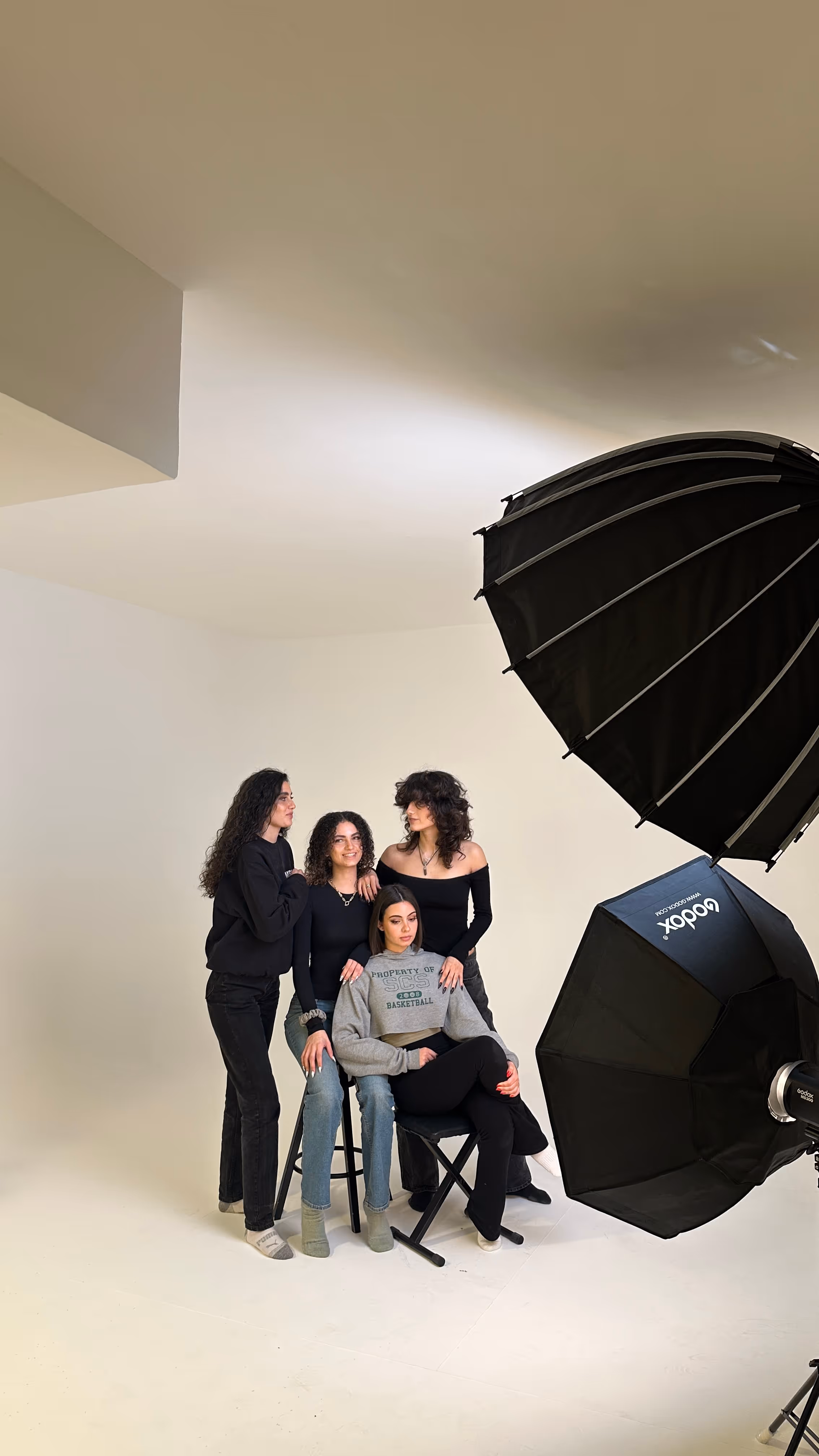 Four women posing in a photo studio with two large black softbox lights visible on the right side.