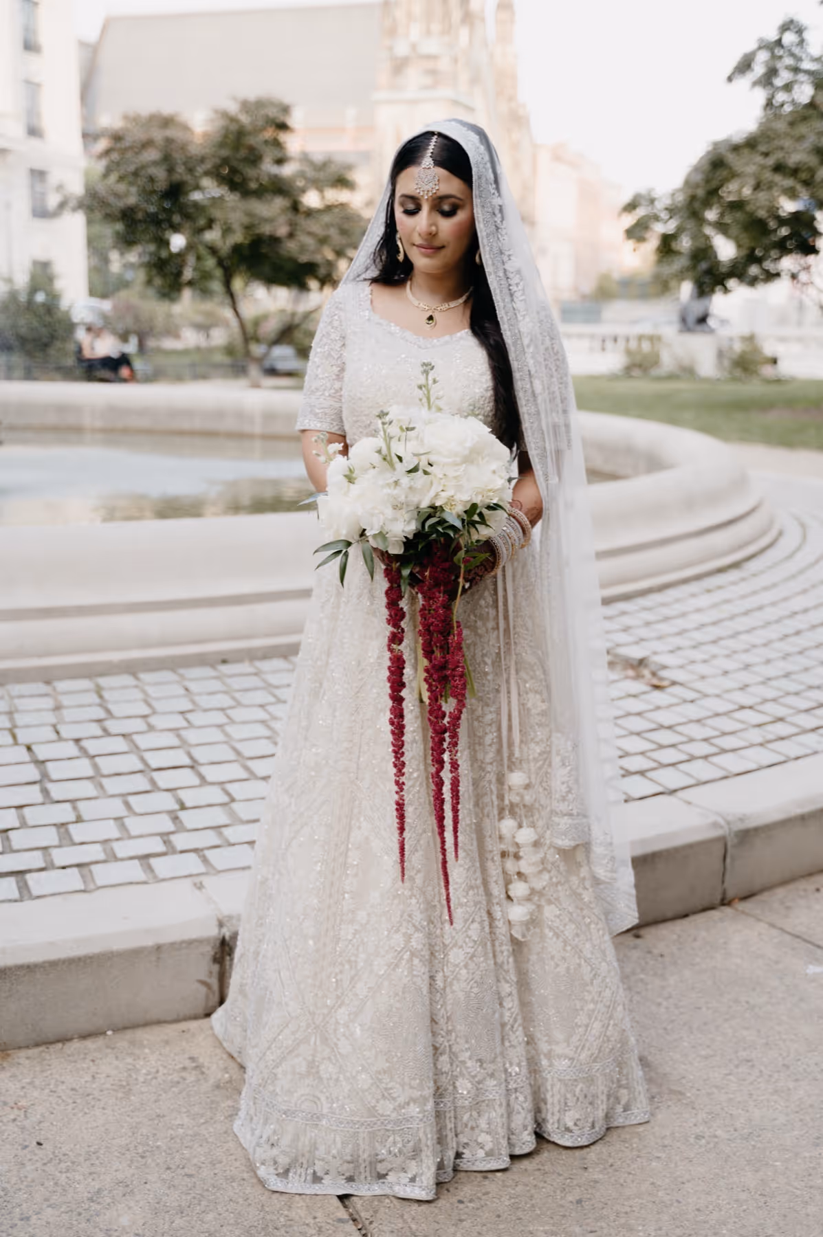 Bride in an intricately embroidered white gown and veil holding a cascading bouquet of white and red flowers outdoors.