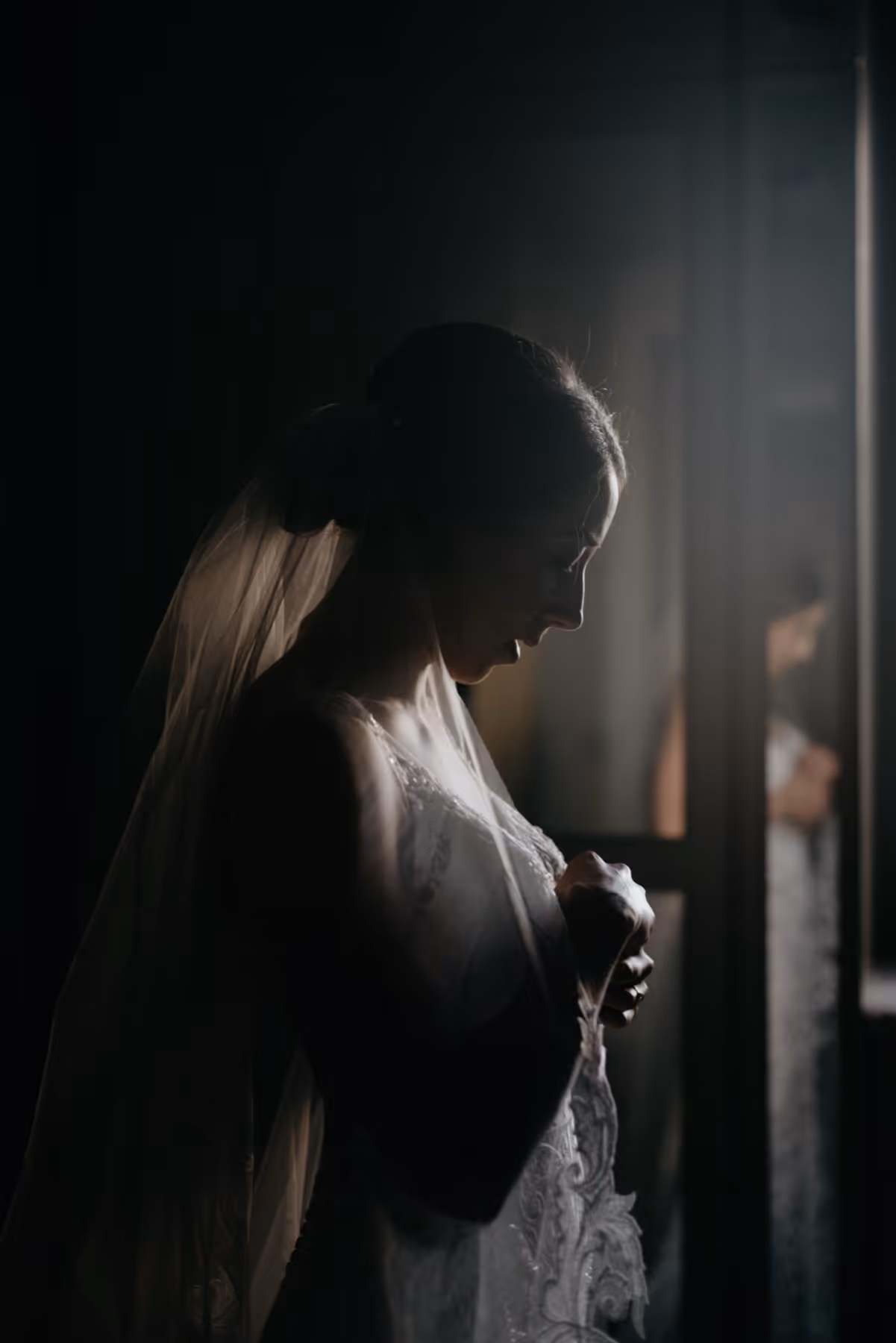 Silhouette of a bride in a wedding dress and veil standing in soft light near a window.