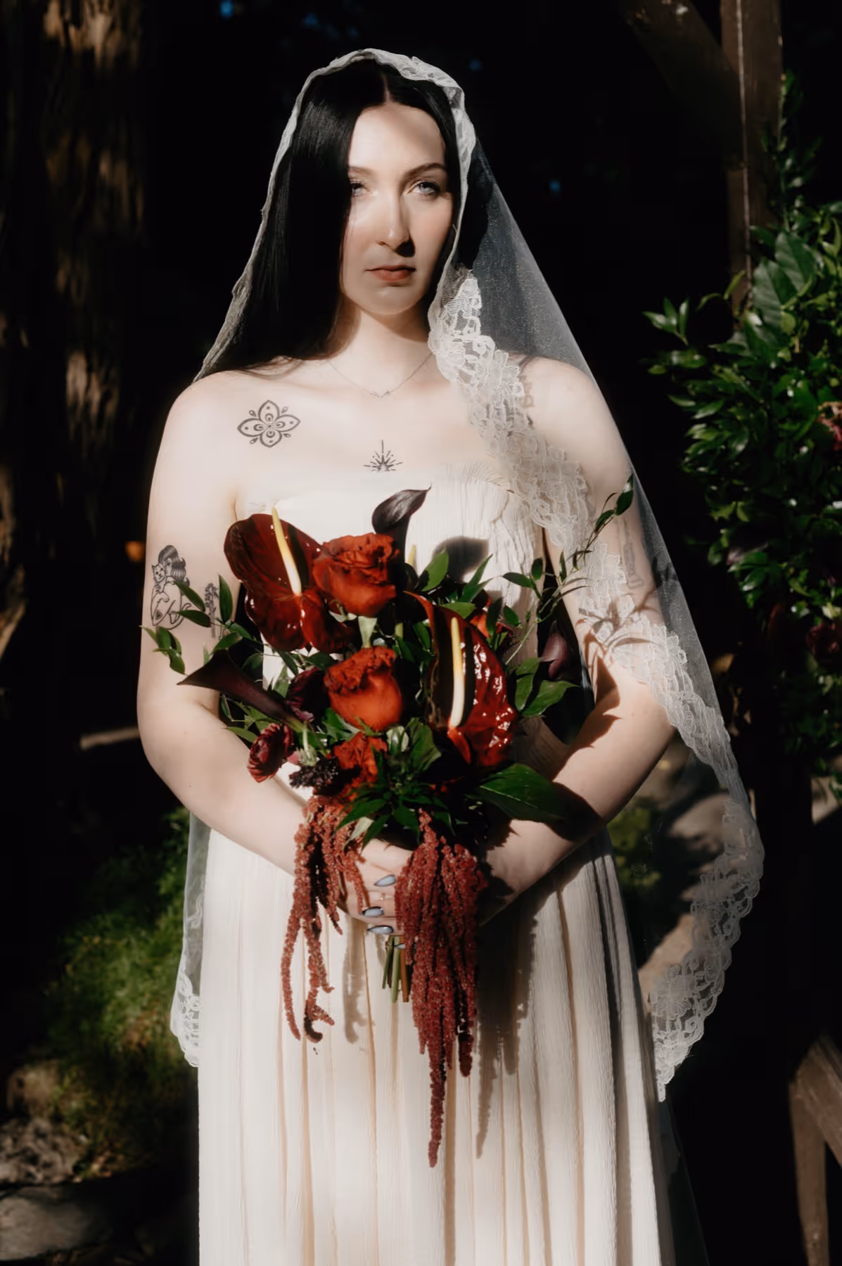Bride with long dark hair wearing a lace veil and sleeveless dress holding a bouquet of red flowers and greenery.