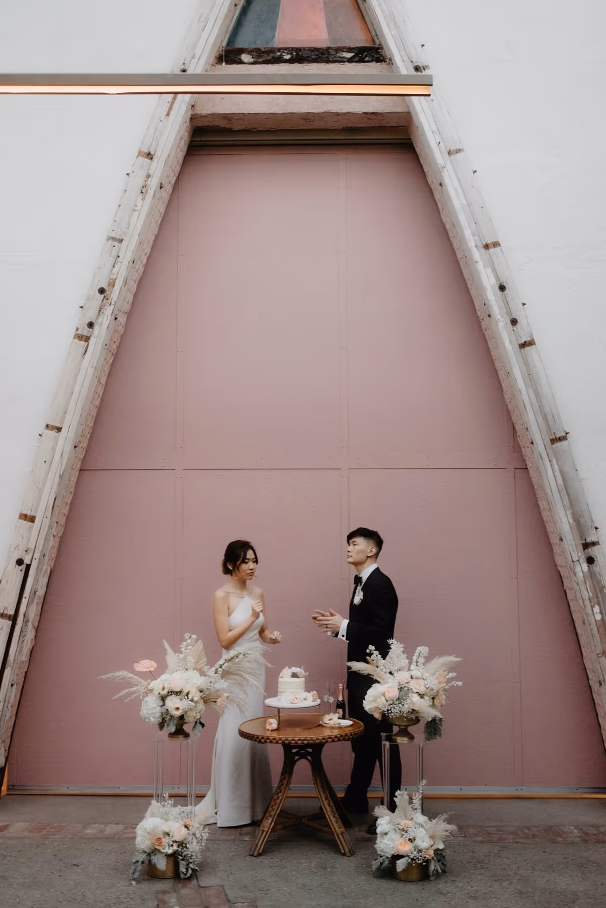 Bride in white dress and groom in black suit standing by a small wooden table with wedding cake and floral arrangements against a large pink triangular door.