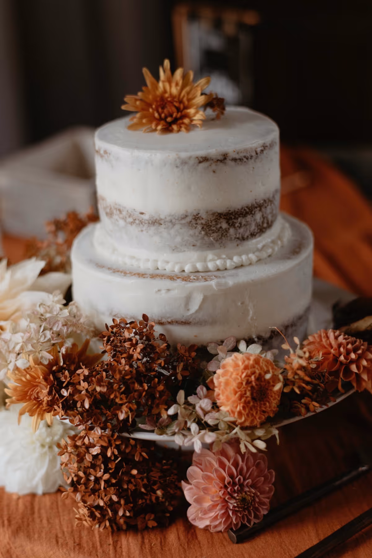 Two-tier rustic wedding cake with white frosting and decorated with dried autumn flowers in orange, brown, and cream tones.