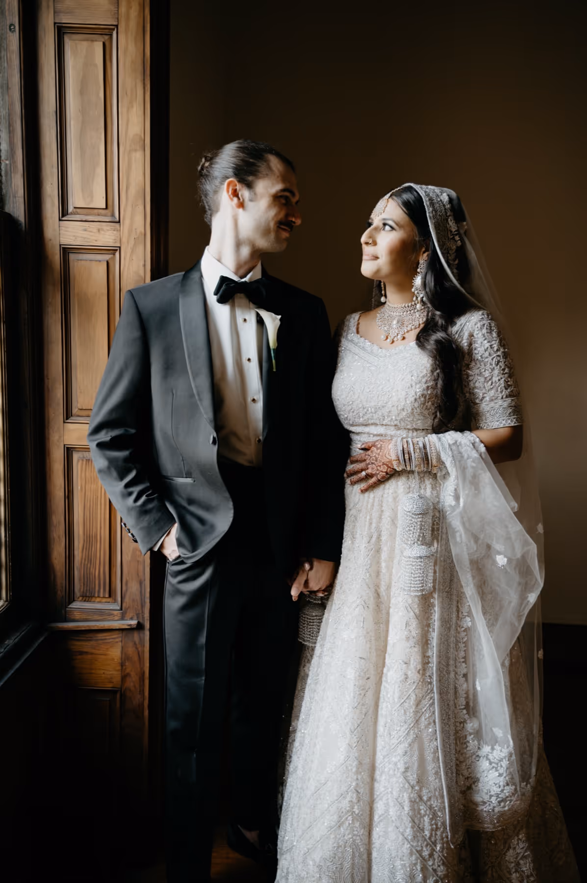 Bride in traditional white embellished South Asian attire with jewelry holding hands with groom in black tuxedo, standing by a wooden window.