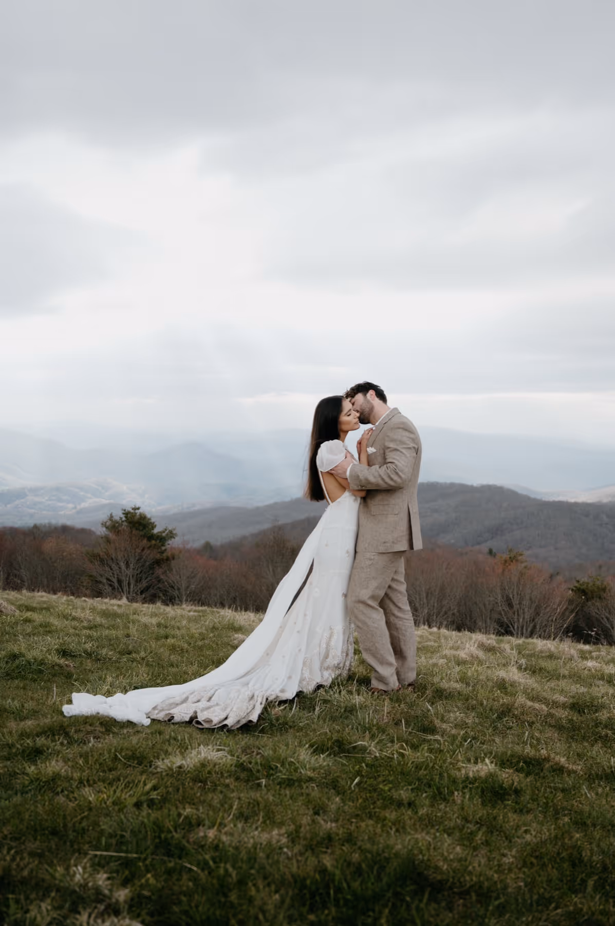 Bride and groom embracing and kissing on a grassy hill with misty mountains in the background.