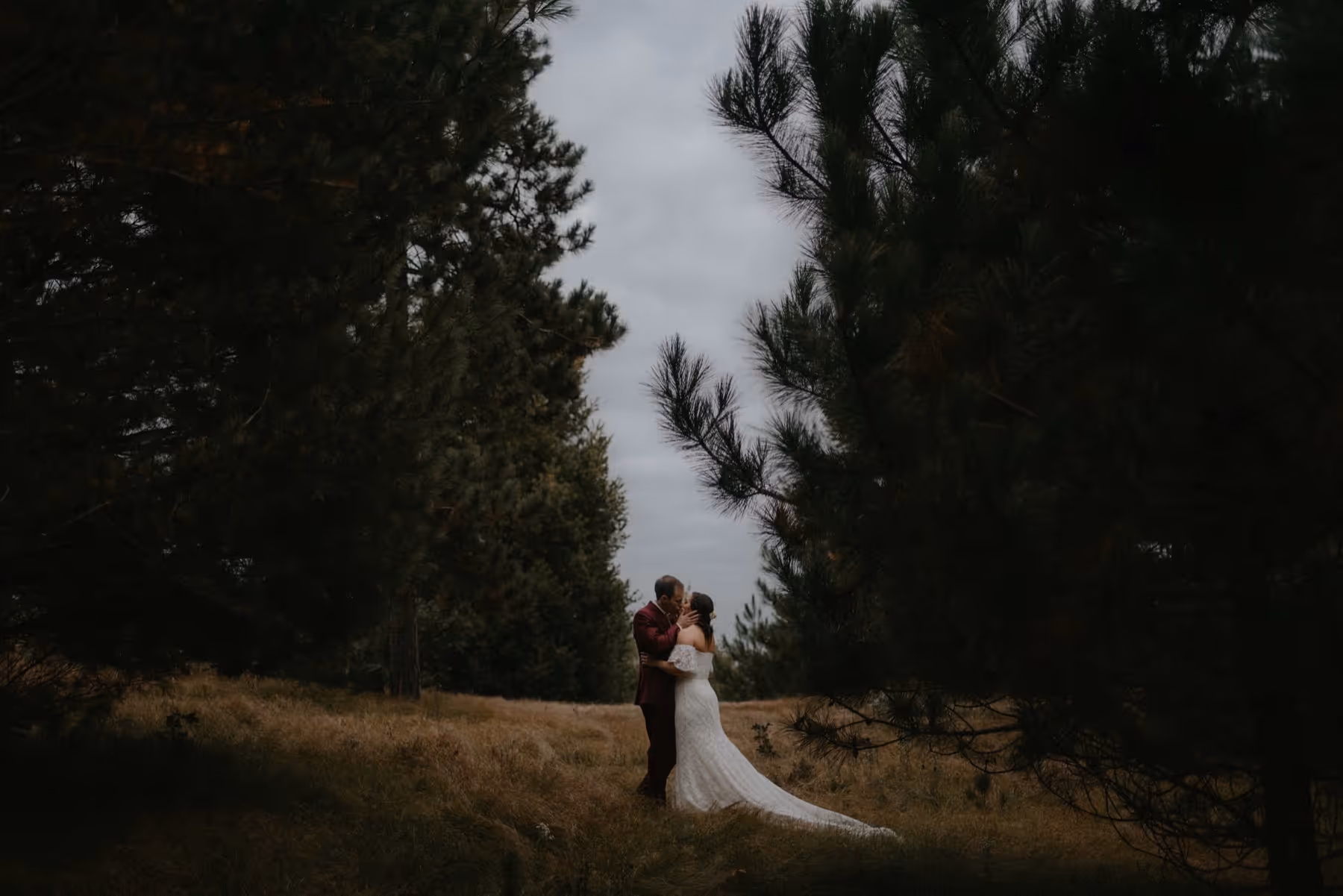 Couple in wedding attire embracing and kissing in a forest clearing surrounded by tall pine trees.