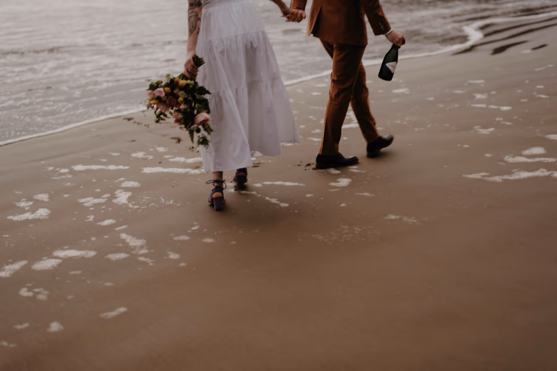 A couple holding hands walking on a beach near the water, the woman holds a bouquet and the man holds a bottle.