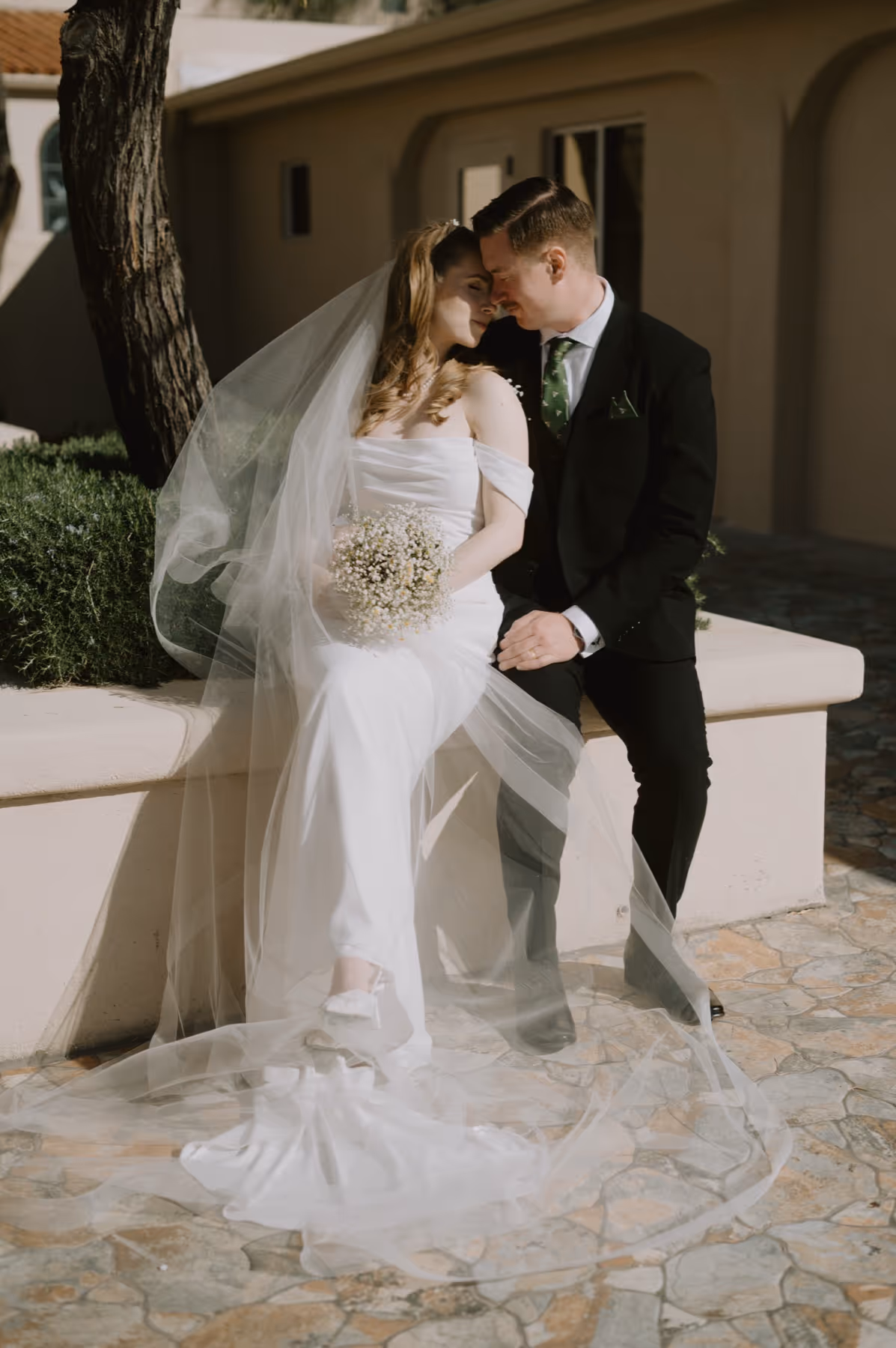 Bride in white dress and veil holding a bouquet sitting next to groom in black suit, both touching foreheads.