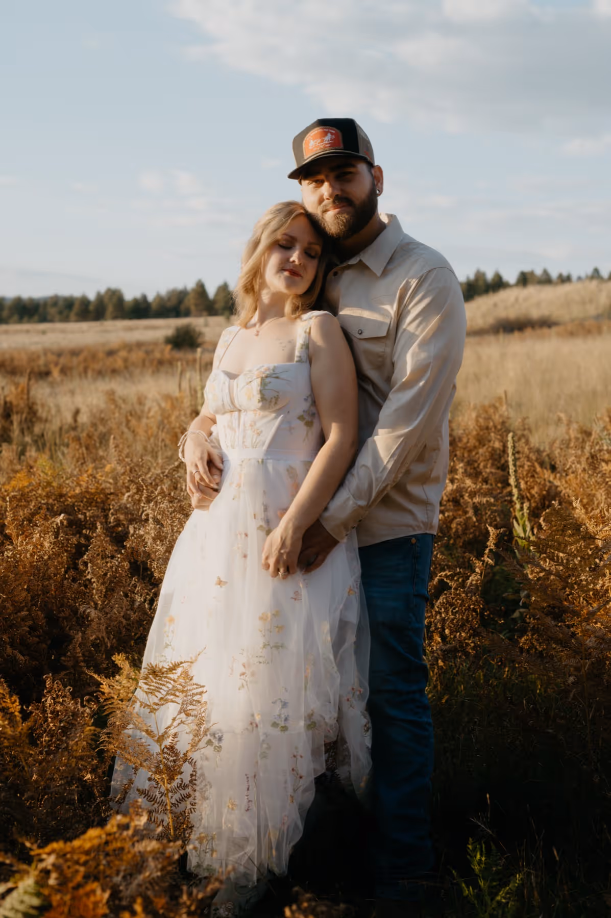 Couple embracing in a field of tall dry plants under a blue sky during golden hour.
