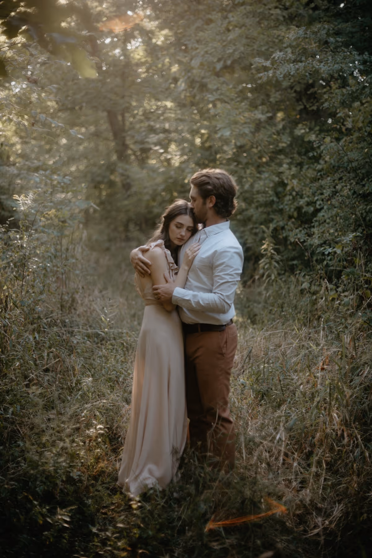 Couple embracing gently in a sunlit forest clearing surrounded by tall grass and trees.