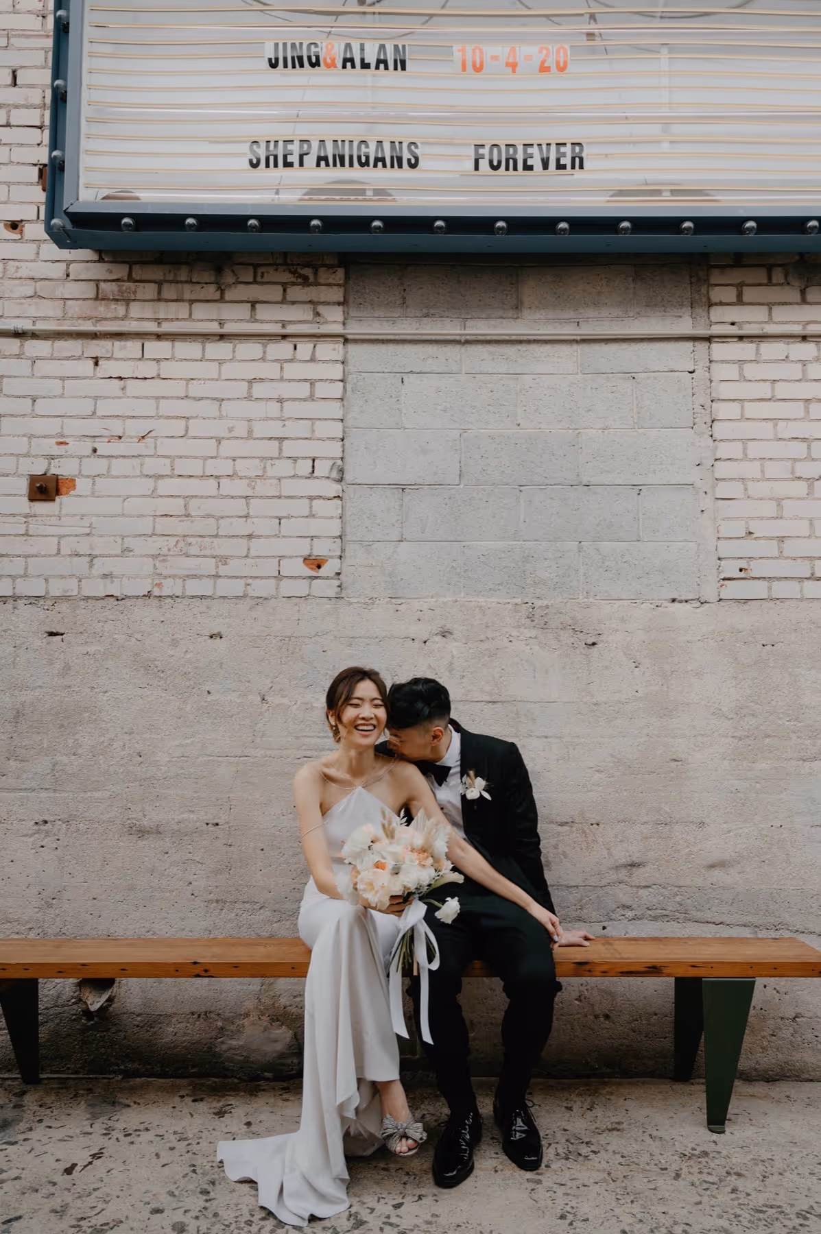 Bride in white dress holding a bouquet and groom in black suit sitting on a bench, smiling and enjoying a moment together under a marquee sign.