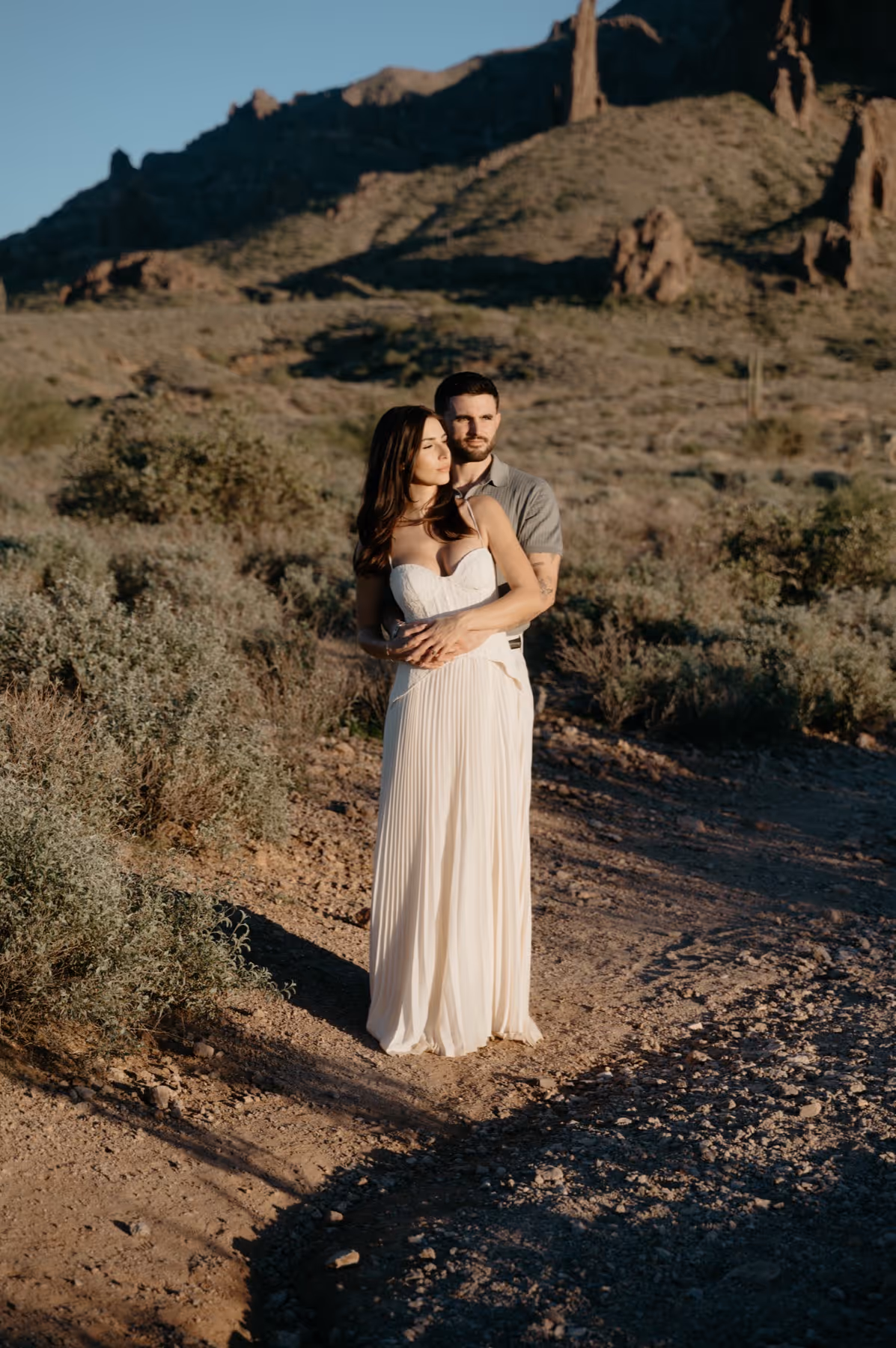 Couple standing in a desert landscape with rocky hills in the background, the woman in a white dress and the man embracing her from behind.