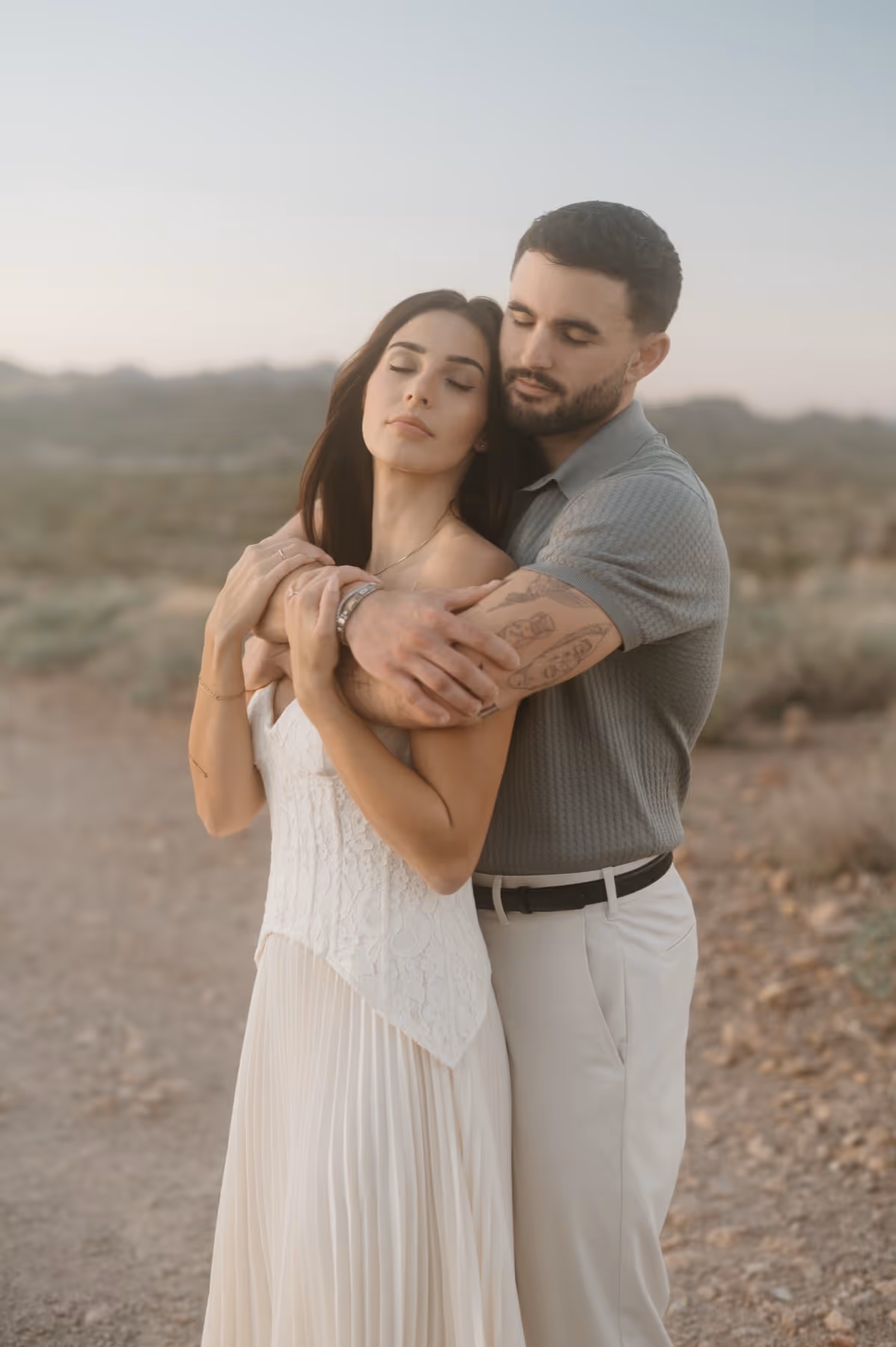 Man with tattoos hugging woman in a white dress in a desert landscape during sunset.