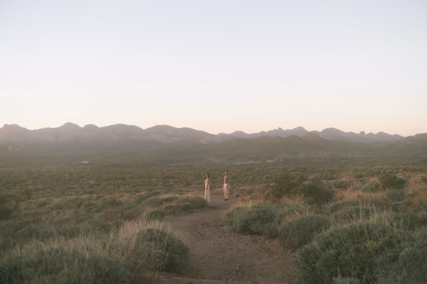 A couple standing on a dirt path in a vast desert landscape with distant mountains under a clear sky at sunset.