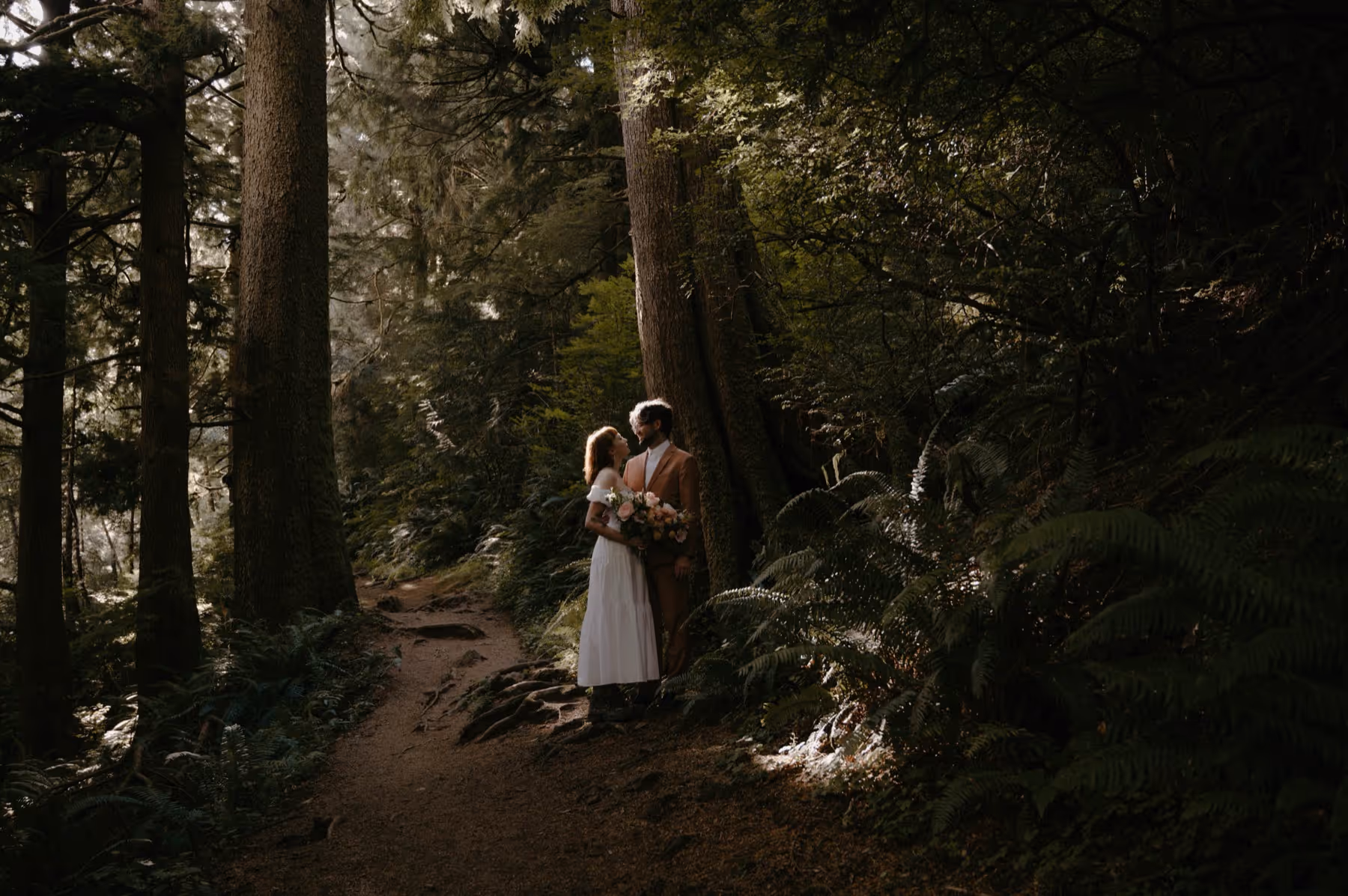 Couple standing closely on a forest path surrounded by tall trees and greenery, holding a bouquet of flowers.