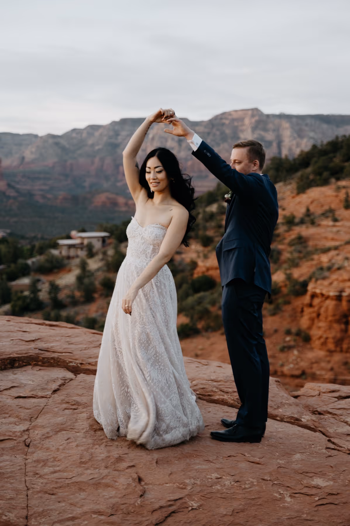 Bride in a white lace dress twirling under groom's raised arm on red rock cliffs at sunset.