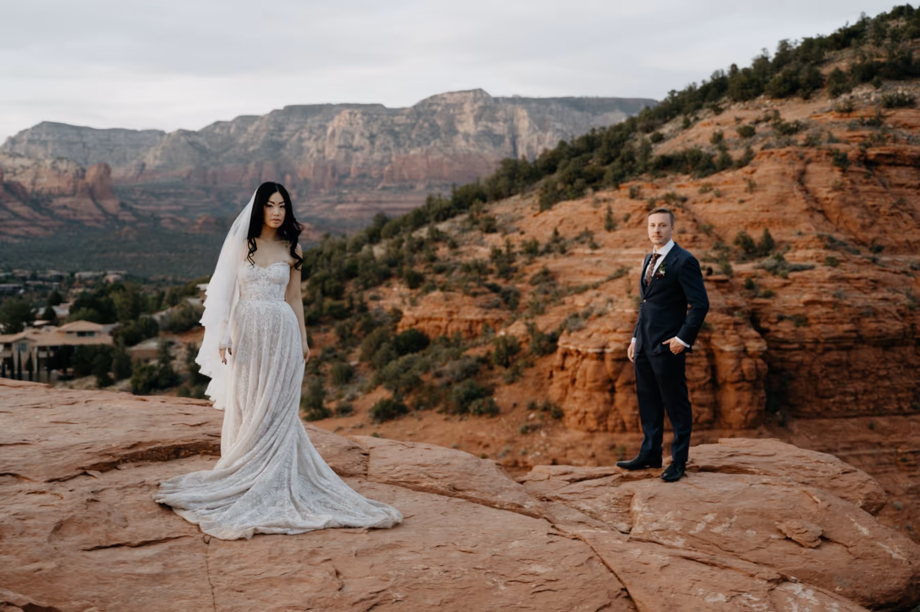 Bride in white wedding dress and veil and groom in dark suit standing on red rock cliffs with mountains in the background.