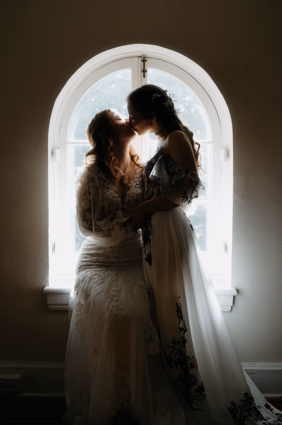 Two women in elegant dresses share a kiss in front of an arched window with bright natural light.