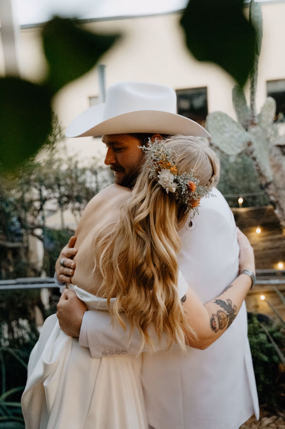 Bride with floral hairpiece and groom in white cowboy hat embracing outdoors.