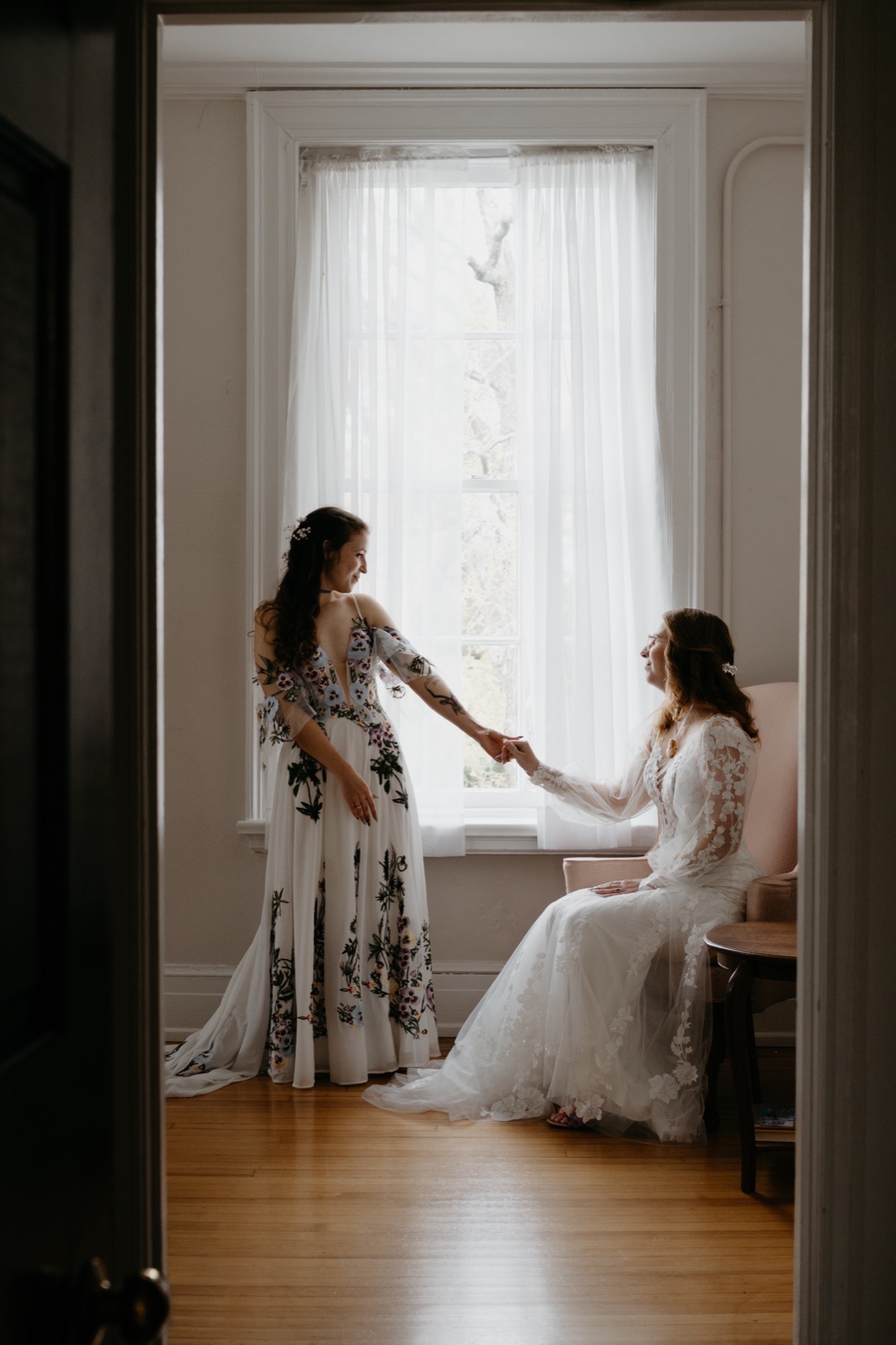 Two women in wedding dresses holding hands near a window with sheer curtains, one standing and one seated in an armchair.