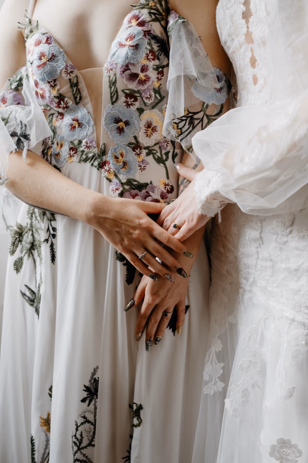 Close-up of two brides wearing floral and lace wedding dresses gently holding hands.