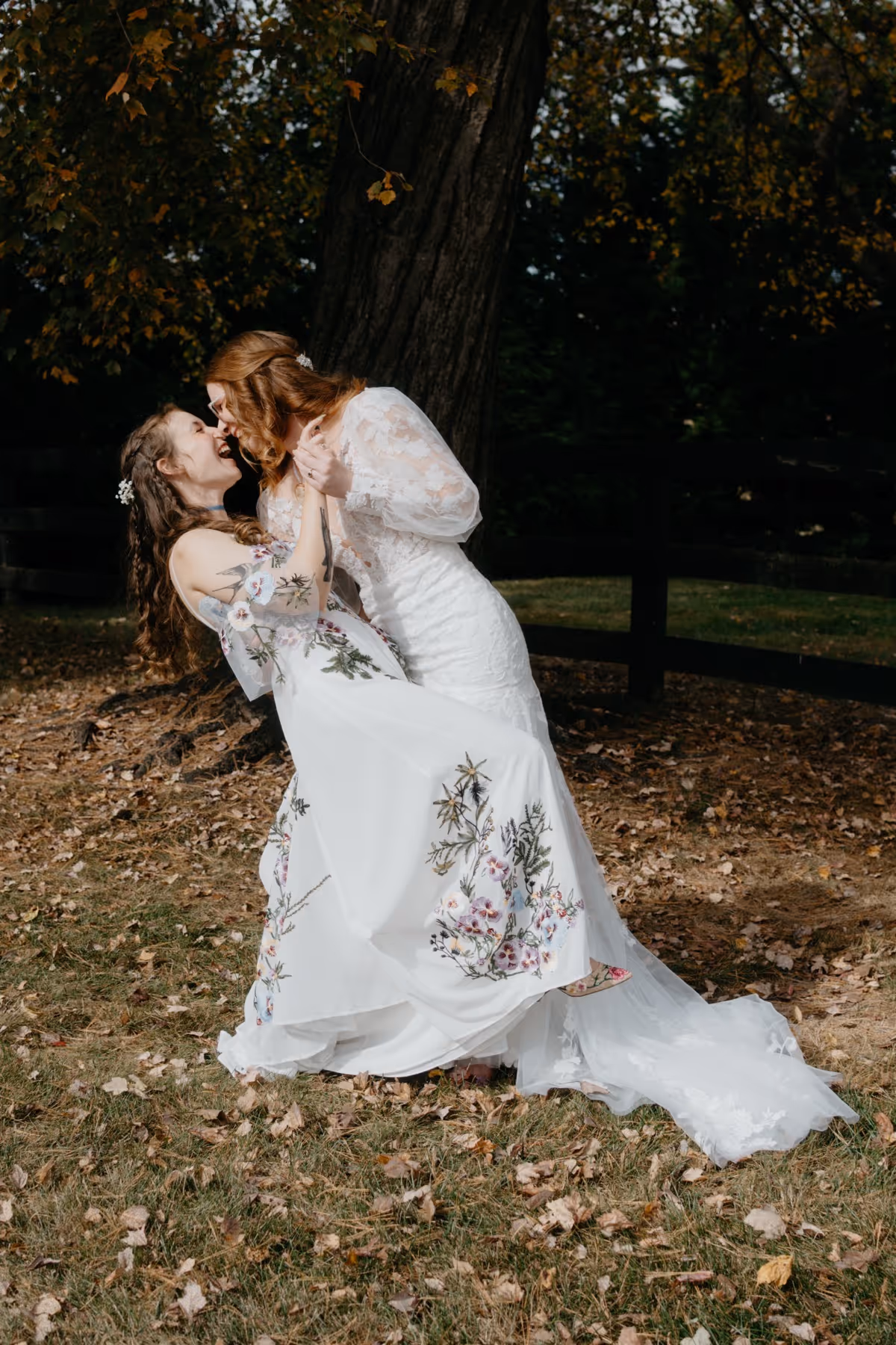 Two brides in wedding dresses share a kiss outdoors near a tree with autumn leaves.