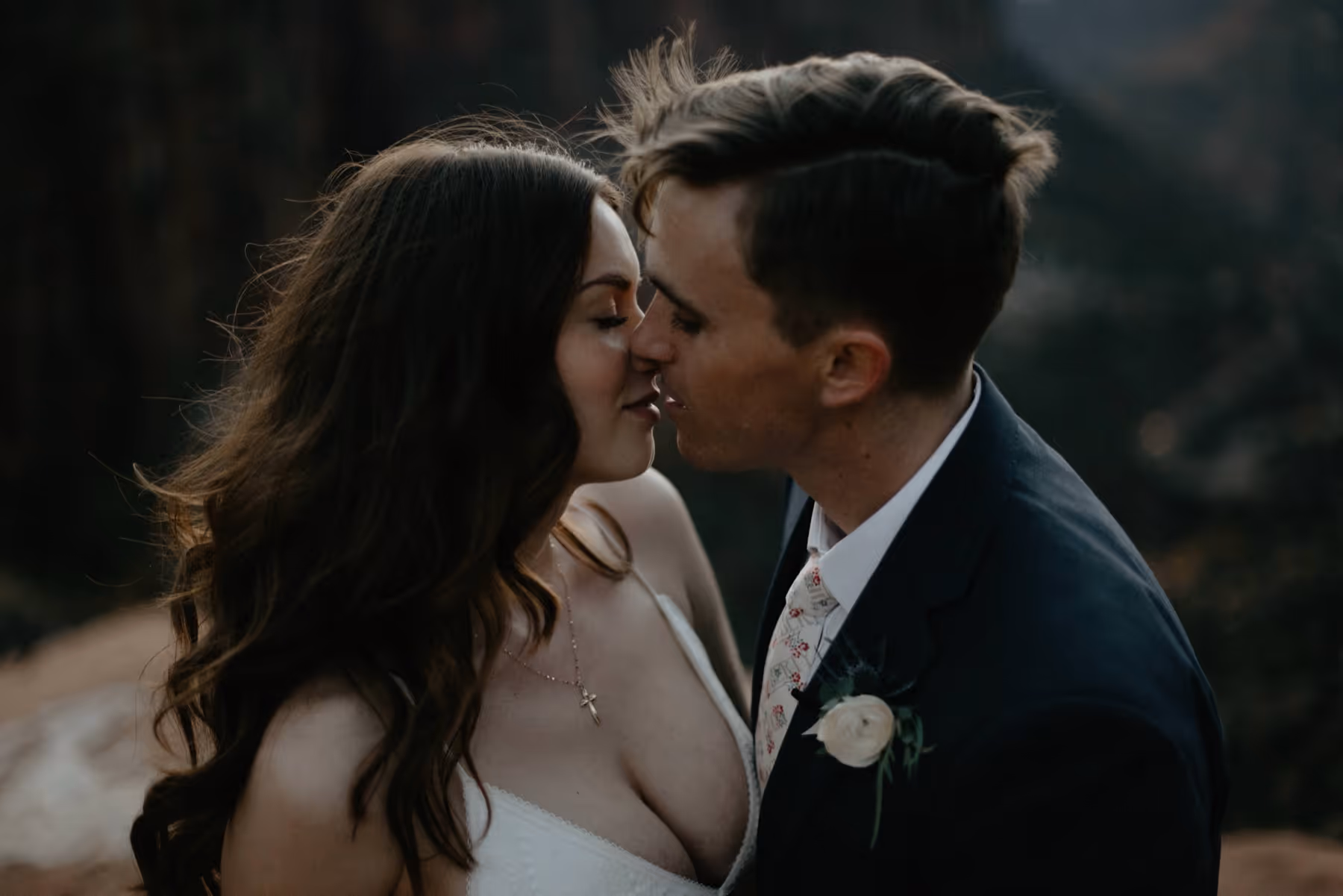 Bride and groom in wedding attire about to kiss with a blurred natural background.