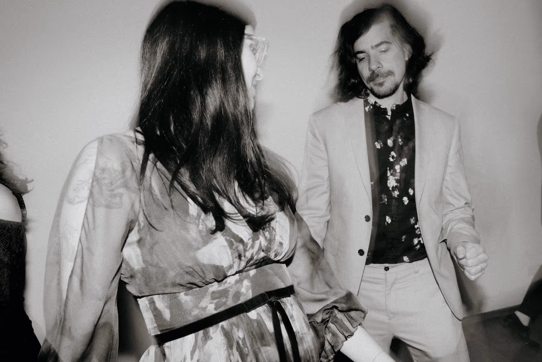 Black and white photo of a woman in a patterned dress dancing with a man in a light suit and floral shirt.